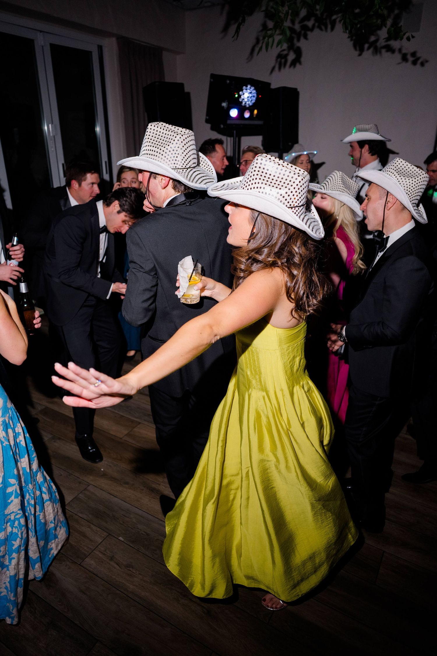 Guests in white cowboy hats dance at a wedding reception, woman in green gown twirls on dance floor.