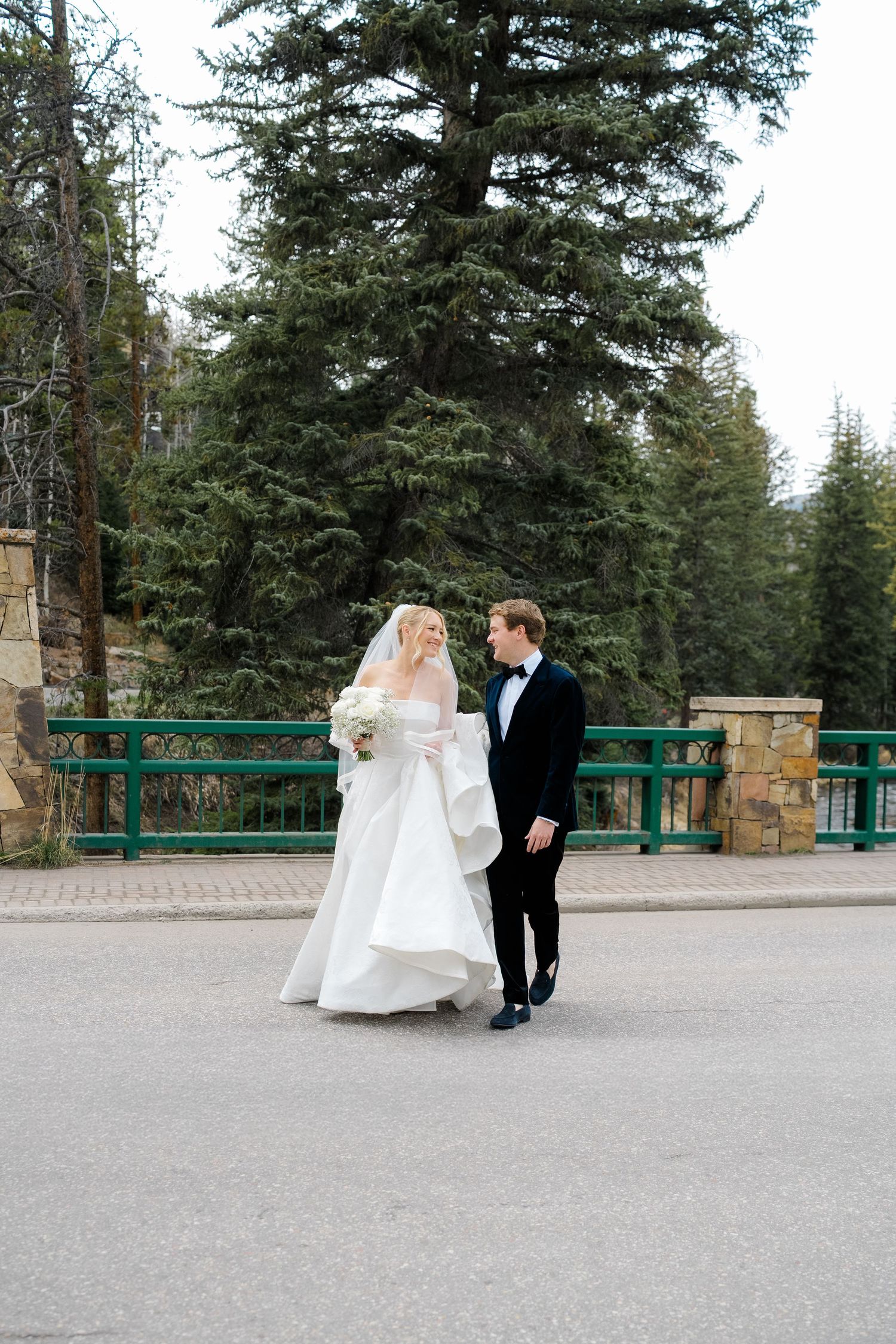 Bride in white gown and groom in tuxedo walking hand in hand on a road surrounded by evergreen trees at Vail Sonnenalp Wedding.