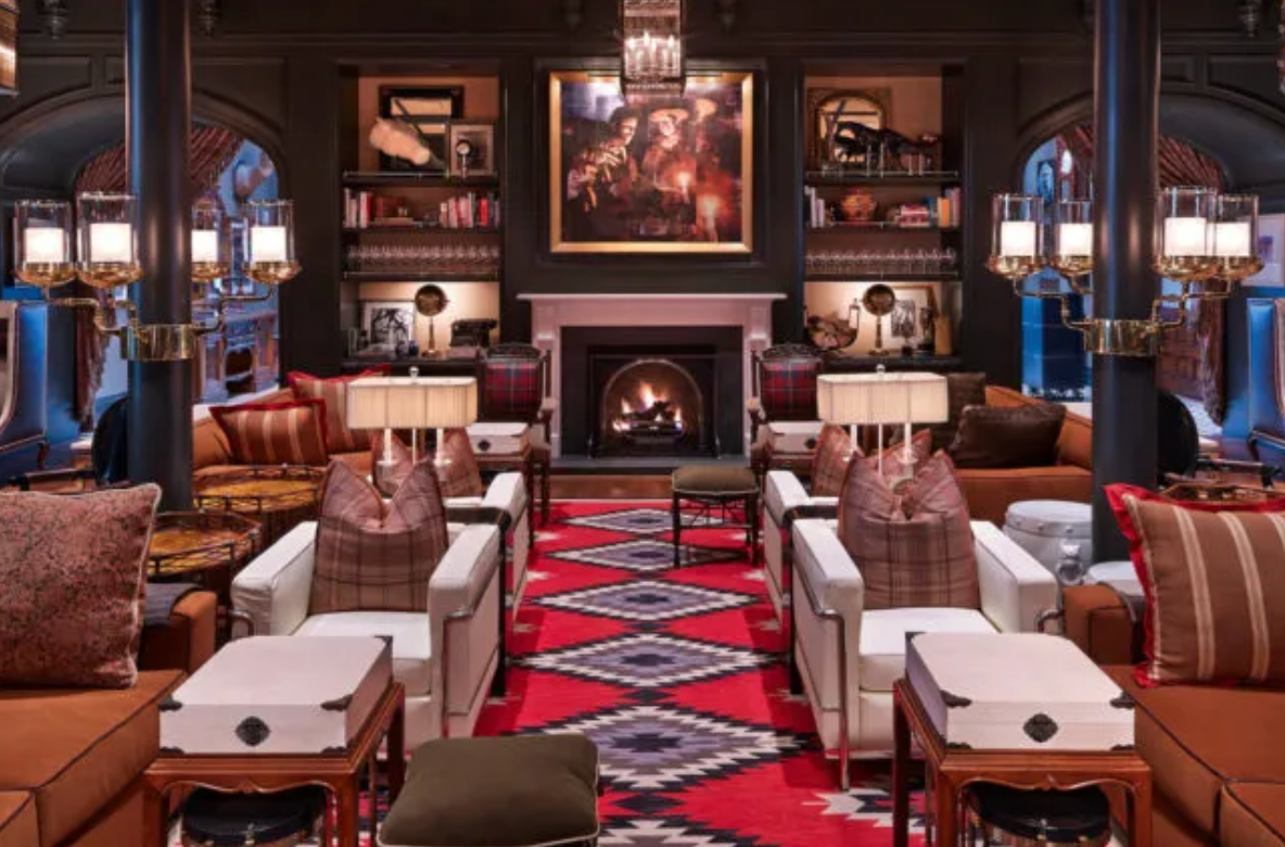 Cozy lodge lounge with red Navajo rug, leather chairs, lit fireplace, warm lighting, and rustic Western decor.