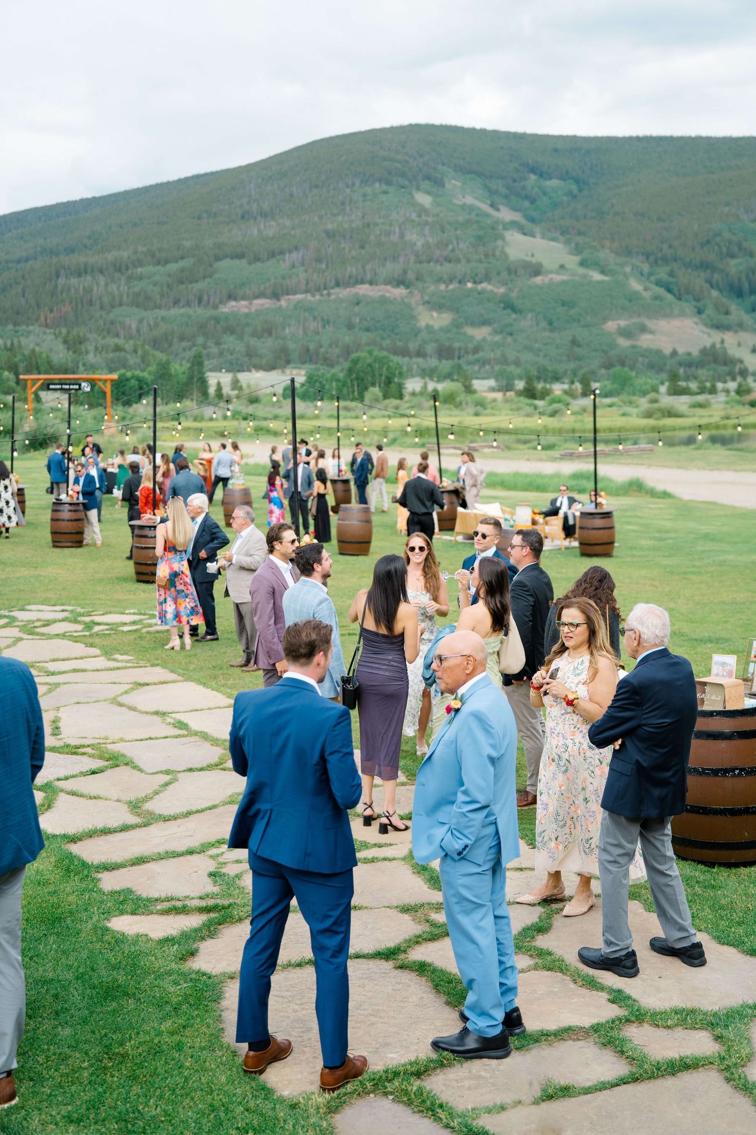 At Camp Hale Wedding Venue -Wedding guests mingle outdoors on stone path with barrel cocktail tables and stunning mountain scenery backdrop.