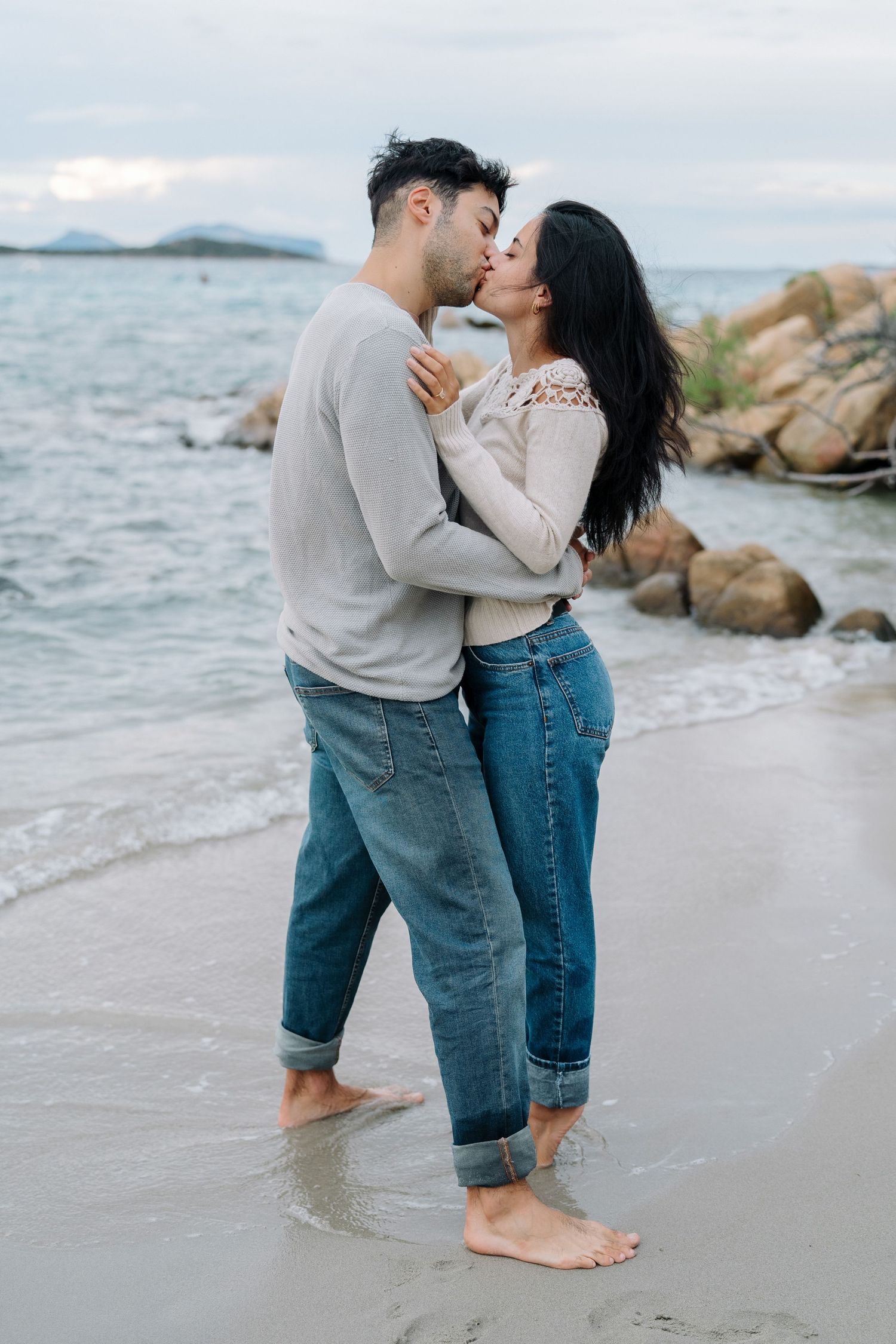 A romantic couple shares tender moments while standing barefoot in the ocean water at sunset along a rocky beach.