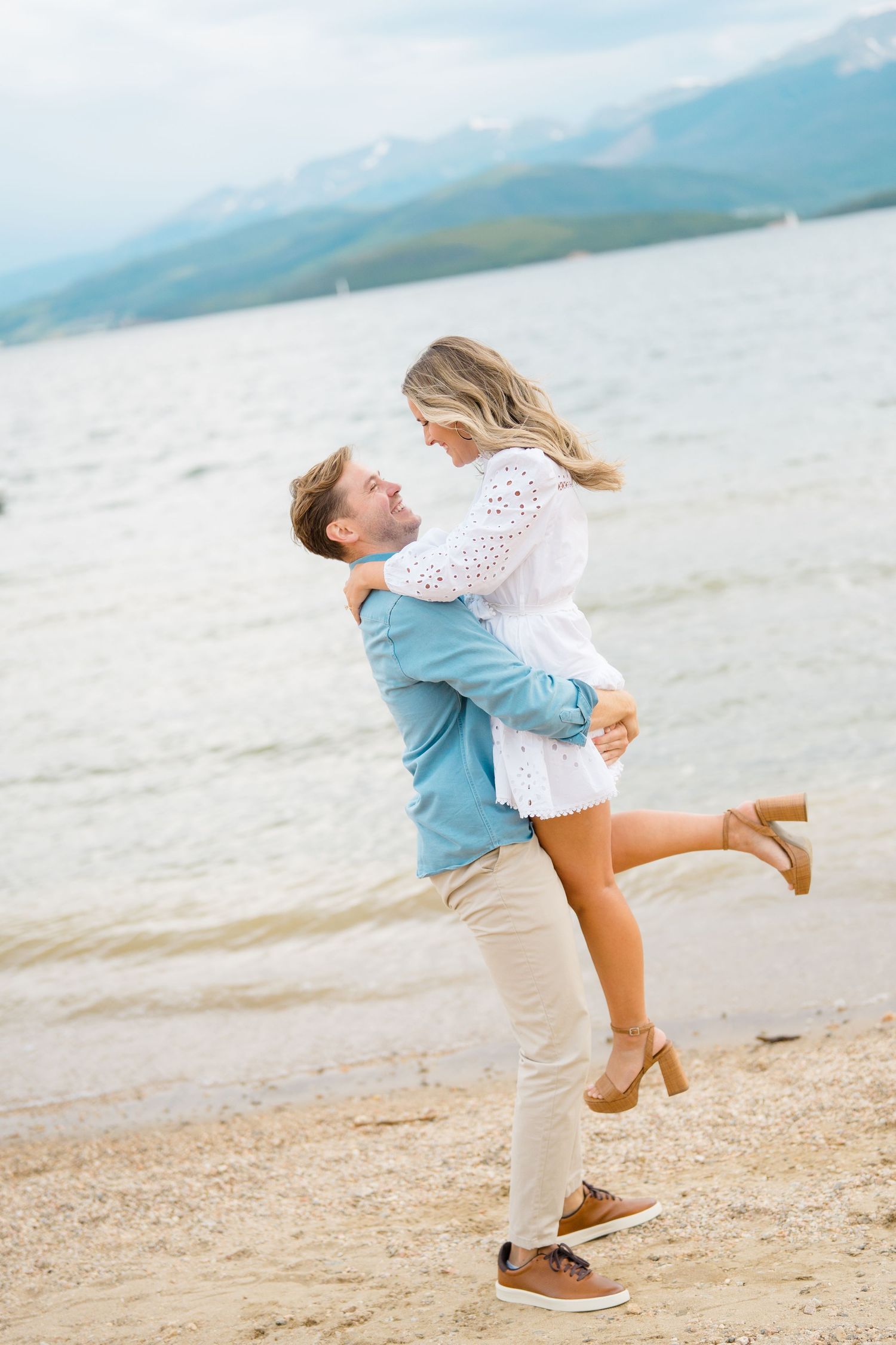 A couple embraces playfully on a beach with mountains and water in the background.