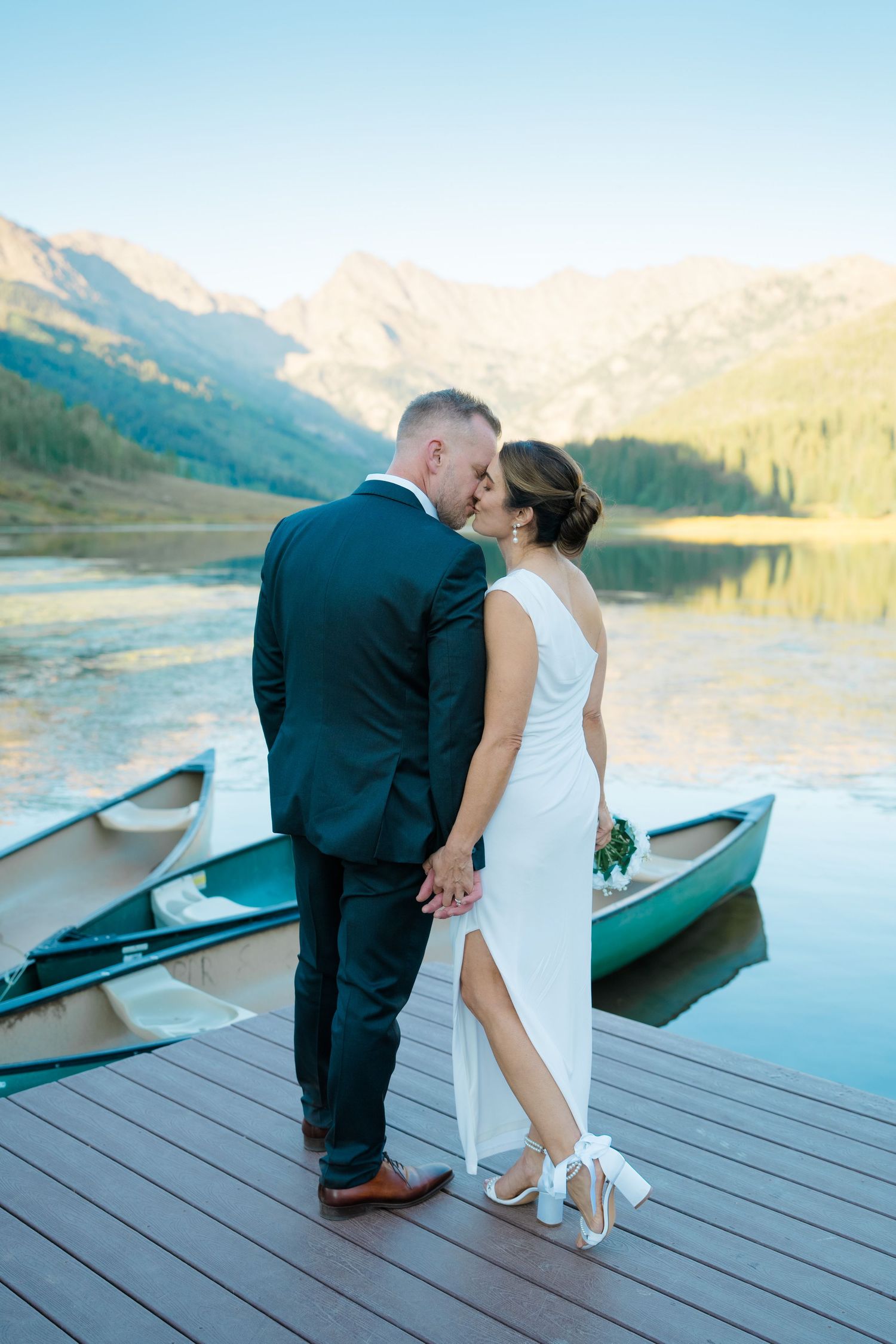 A romantic wedding couple shares intimate moments on a wooden dock by a serene mountain lake with canoes at sunset during piney river ranch
