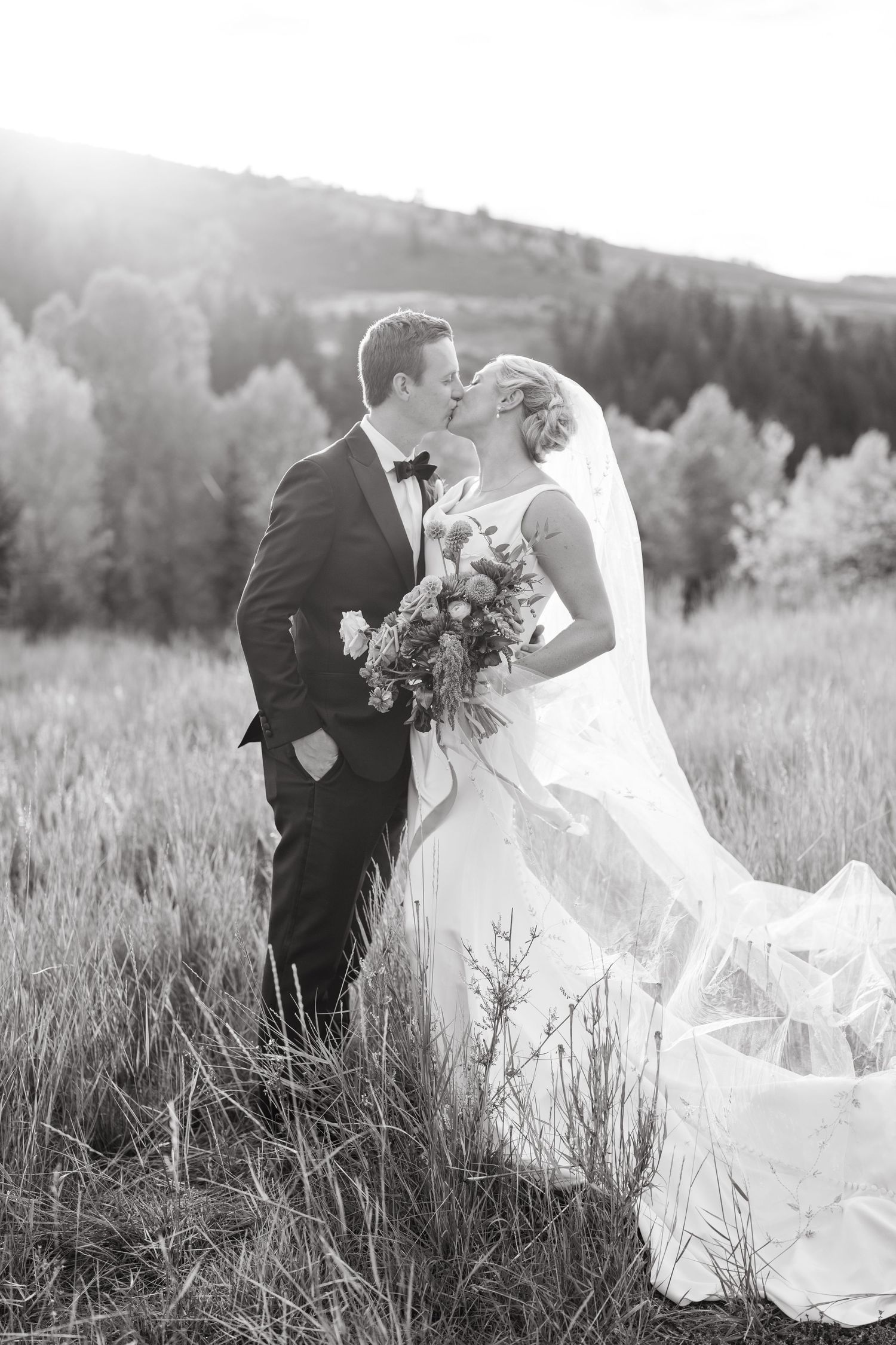 A romantic black and white wedding photo of a couple embracing in a mountain meadow at sunset.