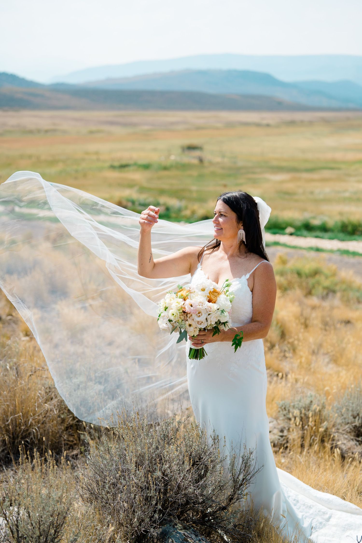 A bride in a white dress holds a flowing veil and bouquet while standing in a scenic mountain landscape at sunset.