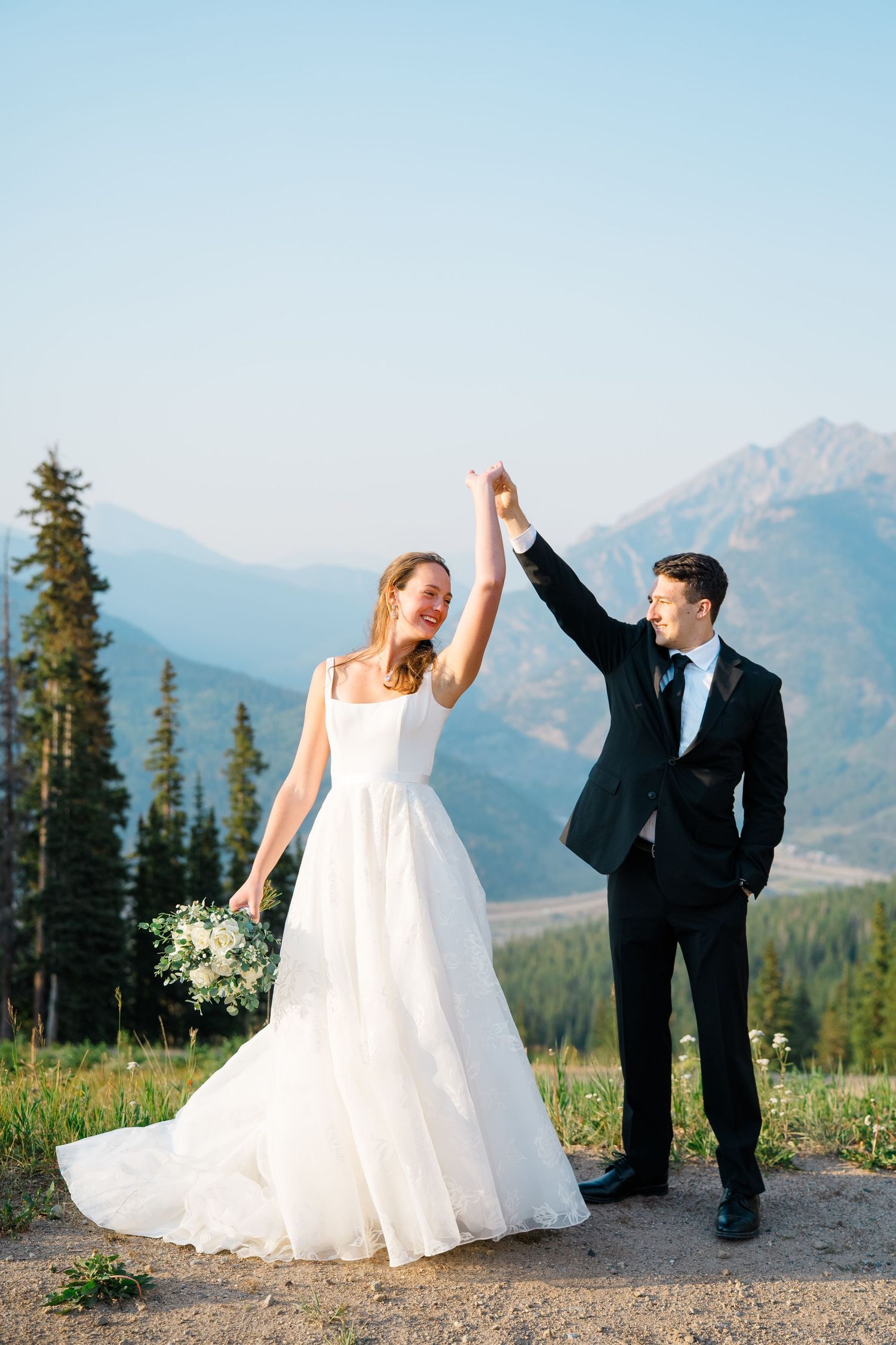 A couple dancing and twirling in formal attire against a scenic mountain backdrop on a sunny day.