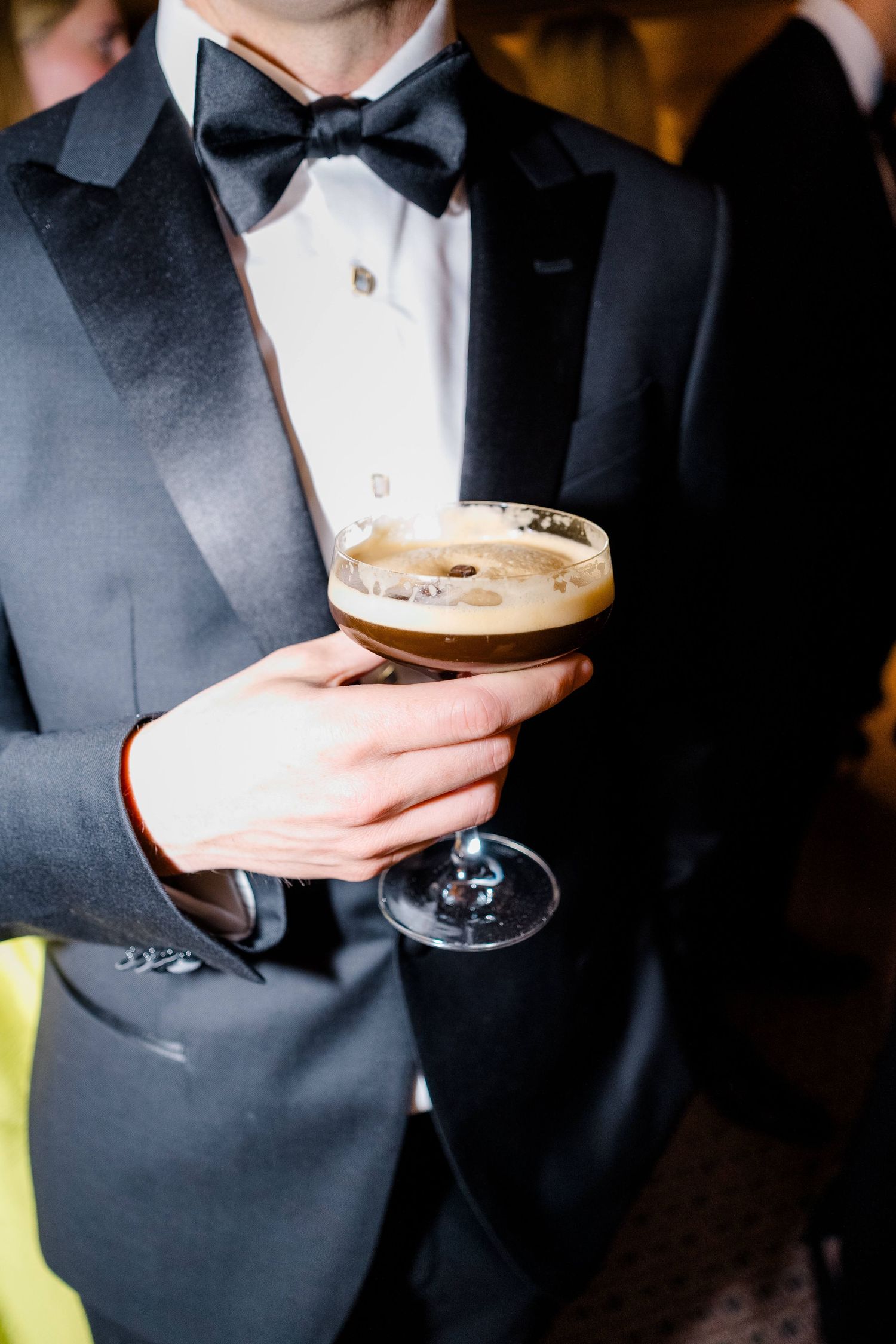 Man in black tuxedo and bow tie holding an espresso martini cocktail in a coupe glass at a formal event.