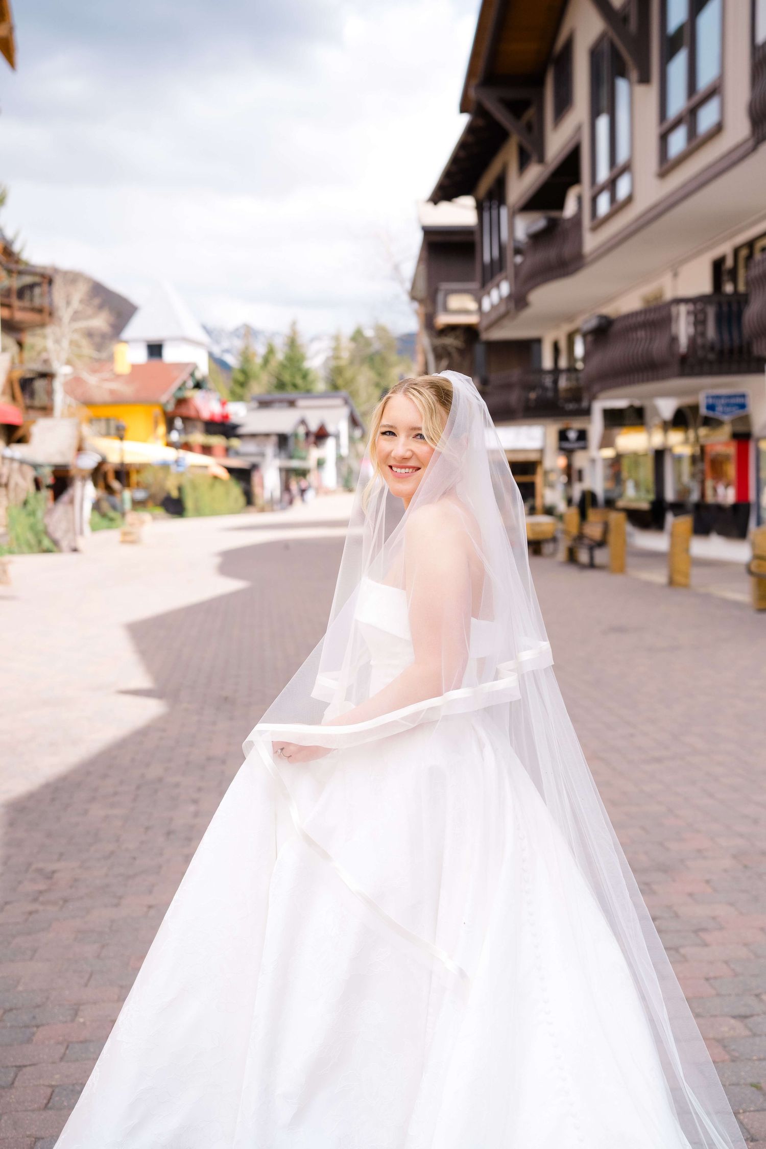Smiling bride in white gown and veil walks through a charming mountain town square on her wedding day.