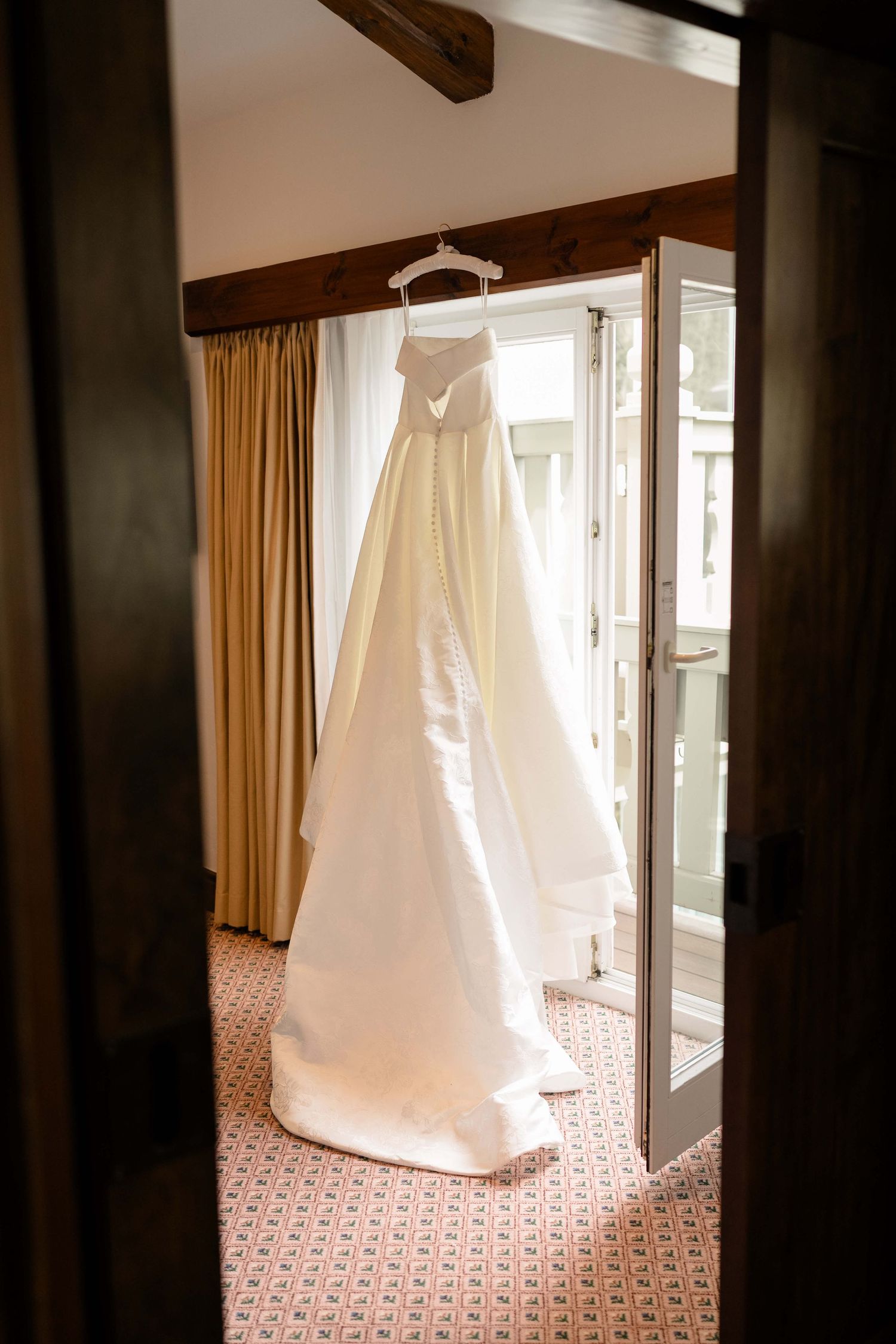 Elegant strapless white wedding gown hanging in a sunlit hotel room doorway at Vail Sonnenalp Wedding.