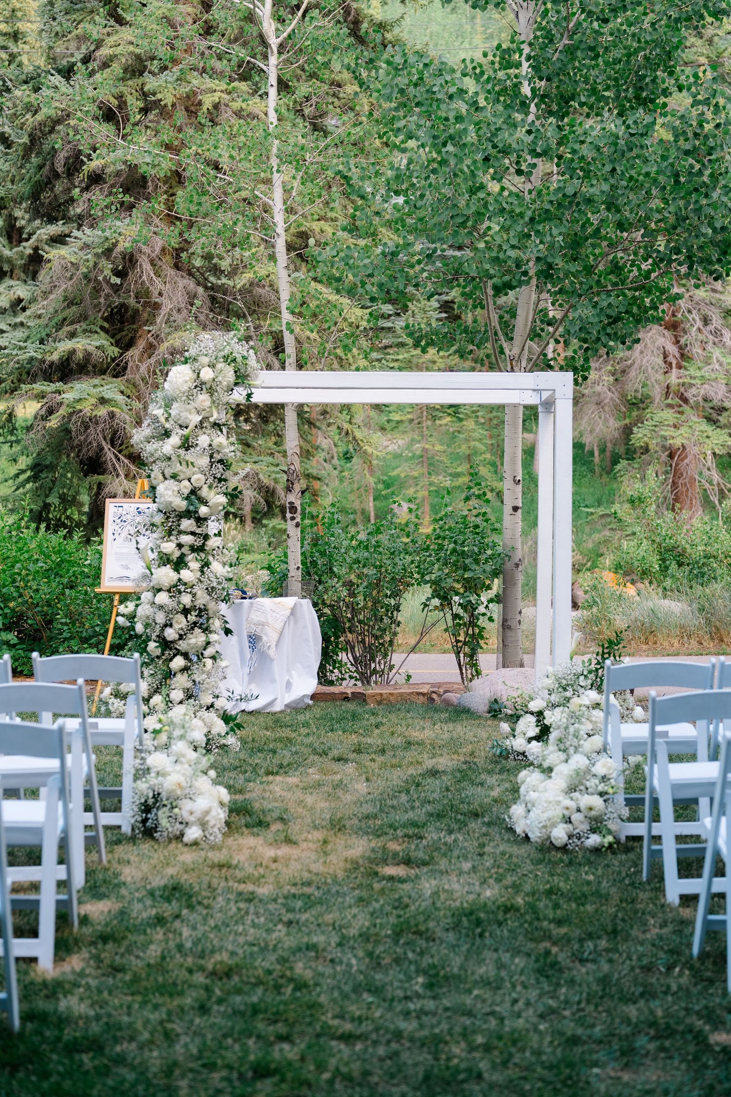 Outdoor wedding ceremony setup with white floral arch, white chairs, and lush green trees in background.