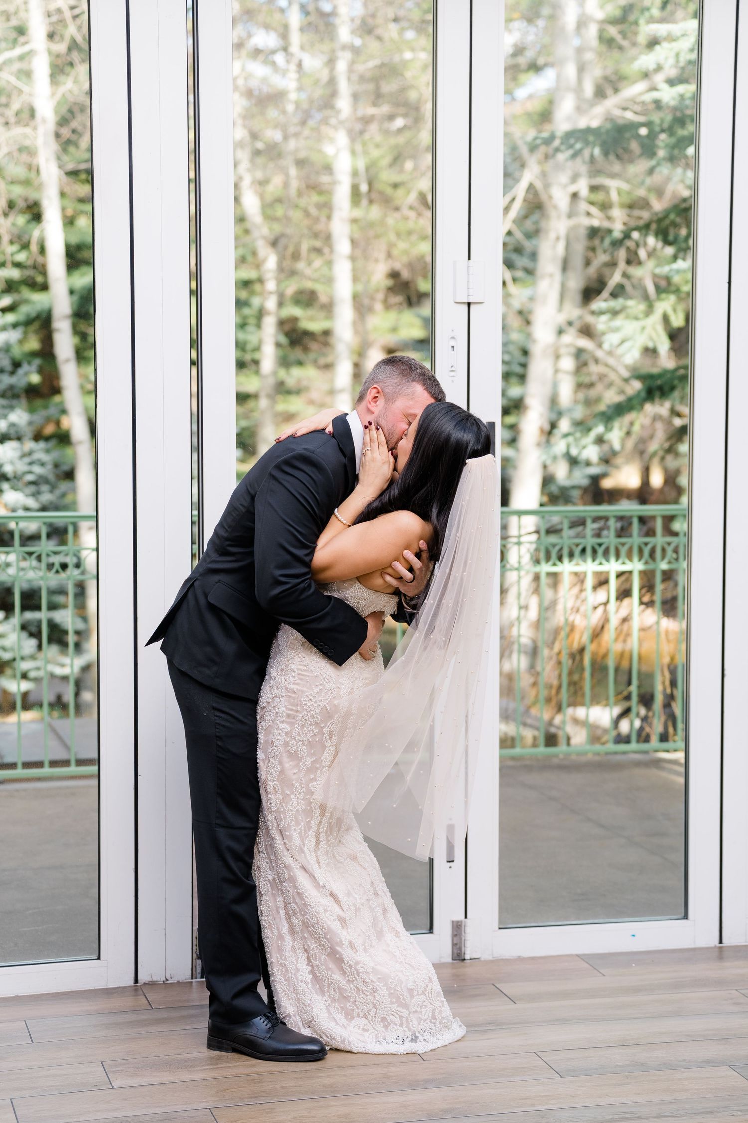 Bride and groom share a romantic kiss in front of glass doors, bride wearing lace gown and veil.