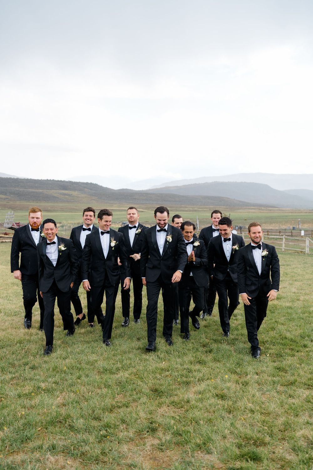 Groom and groomsmen in black tuxedos and bow ties walk together across a grassy field with rolling hills and a cloudy sky behind them.