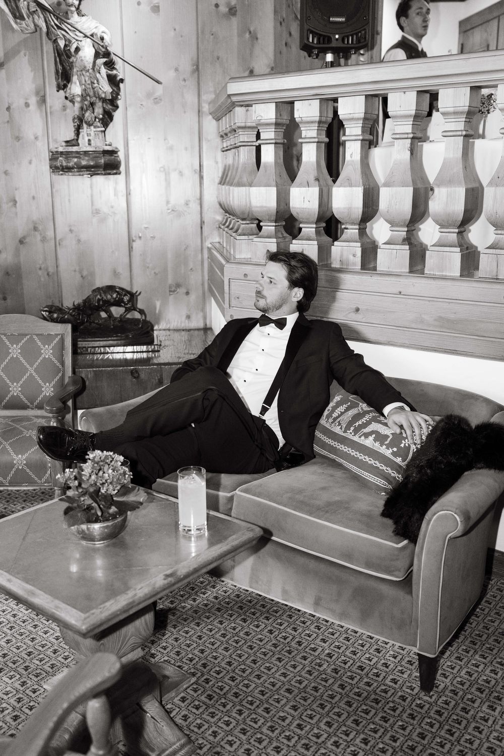 Man in tuxedo relaxing on velvet sofa in elegant wood-paneled room, black and white photo.