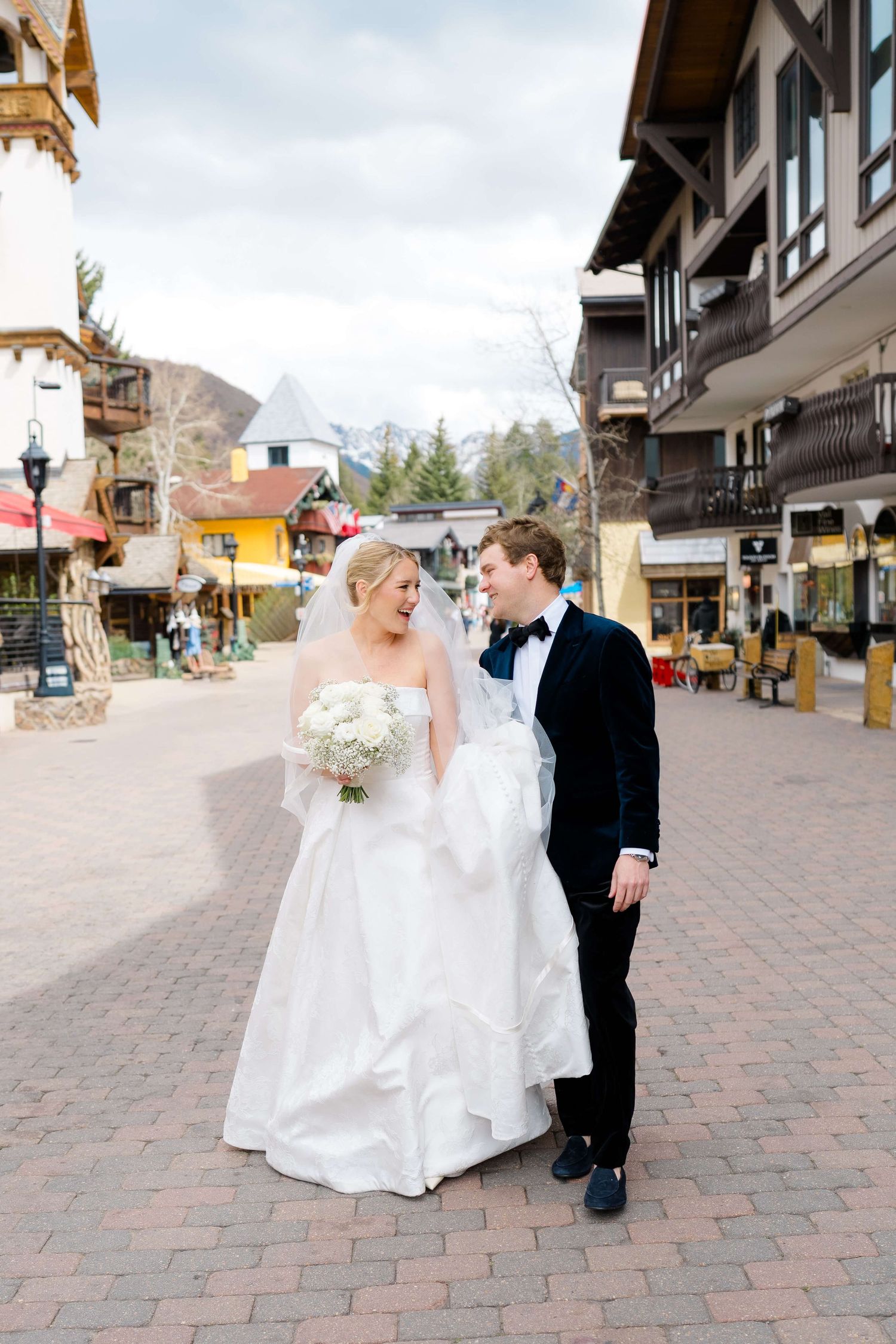 Bride and groom walk through a charming mountain village street, she holds white bouquet, wearing veil and ball gown.