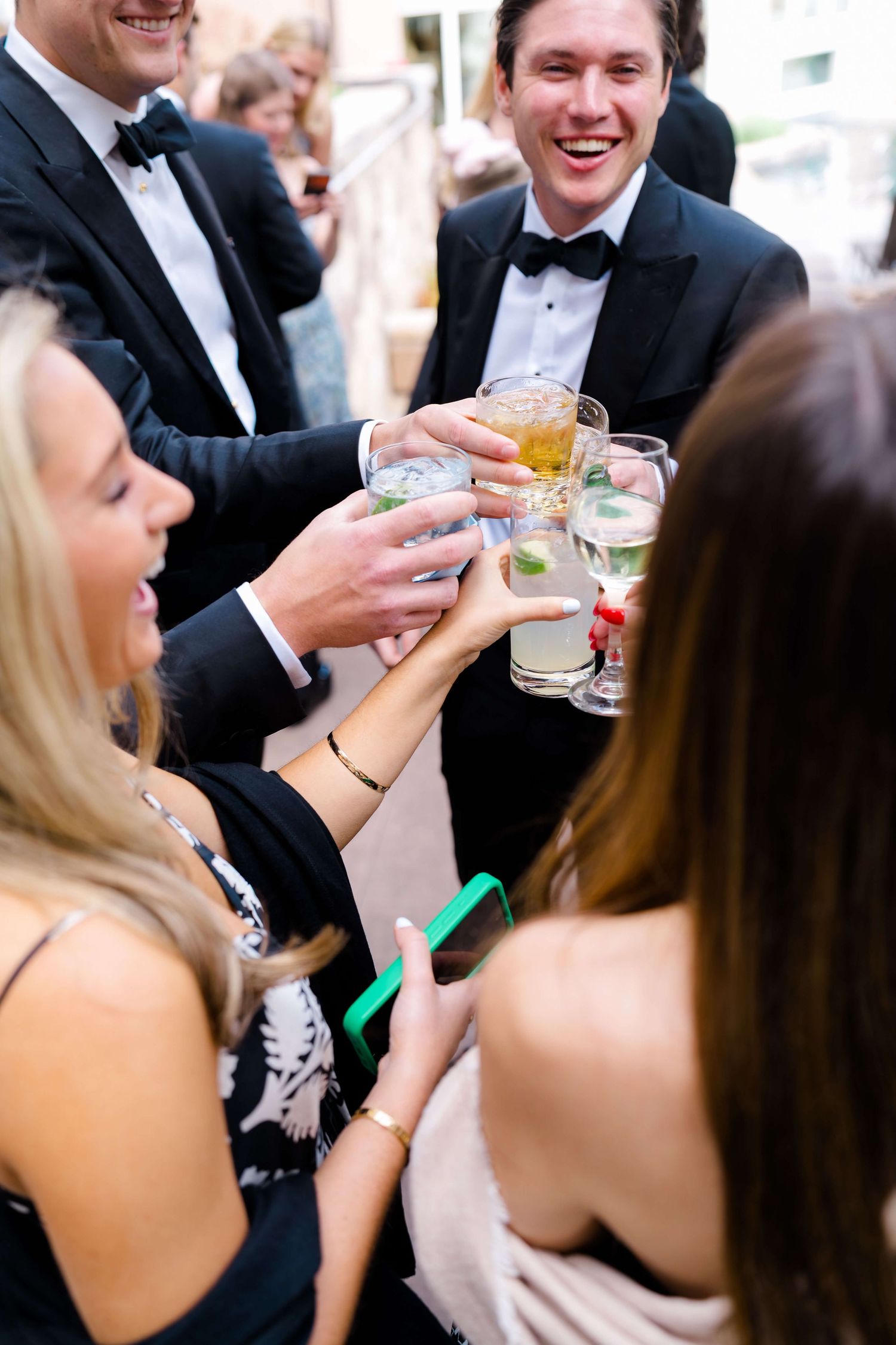 Elegantly dressed guests in tuxedos and formal wear clinking cocktail glasses at an upscale evening event.