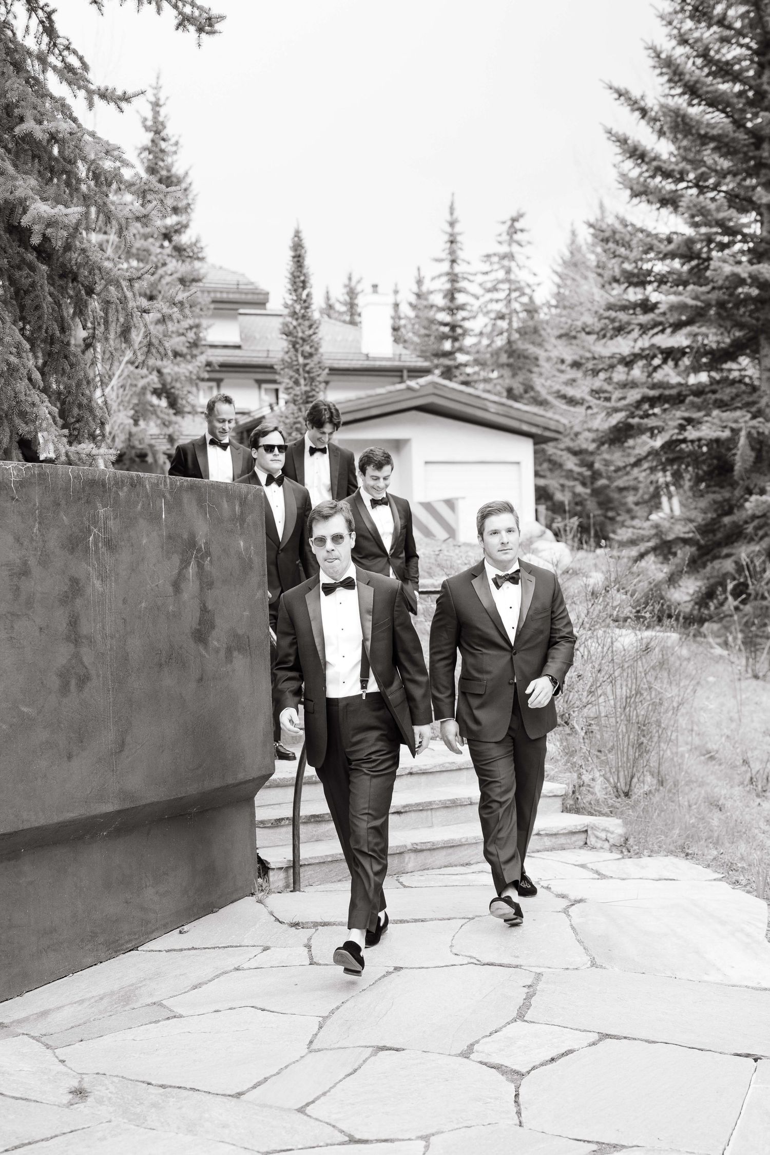 Black and white photo of groomsmen in tuxedos walking confidently along a stone path surrounded by tall evergreen trees.