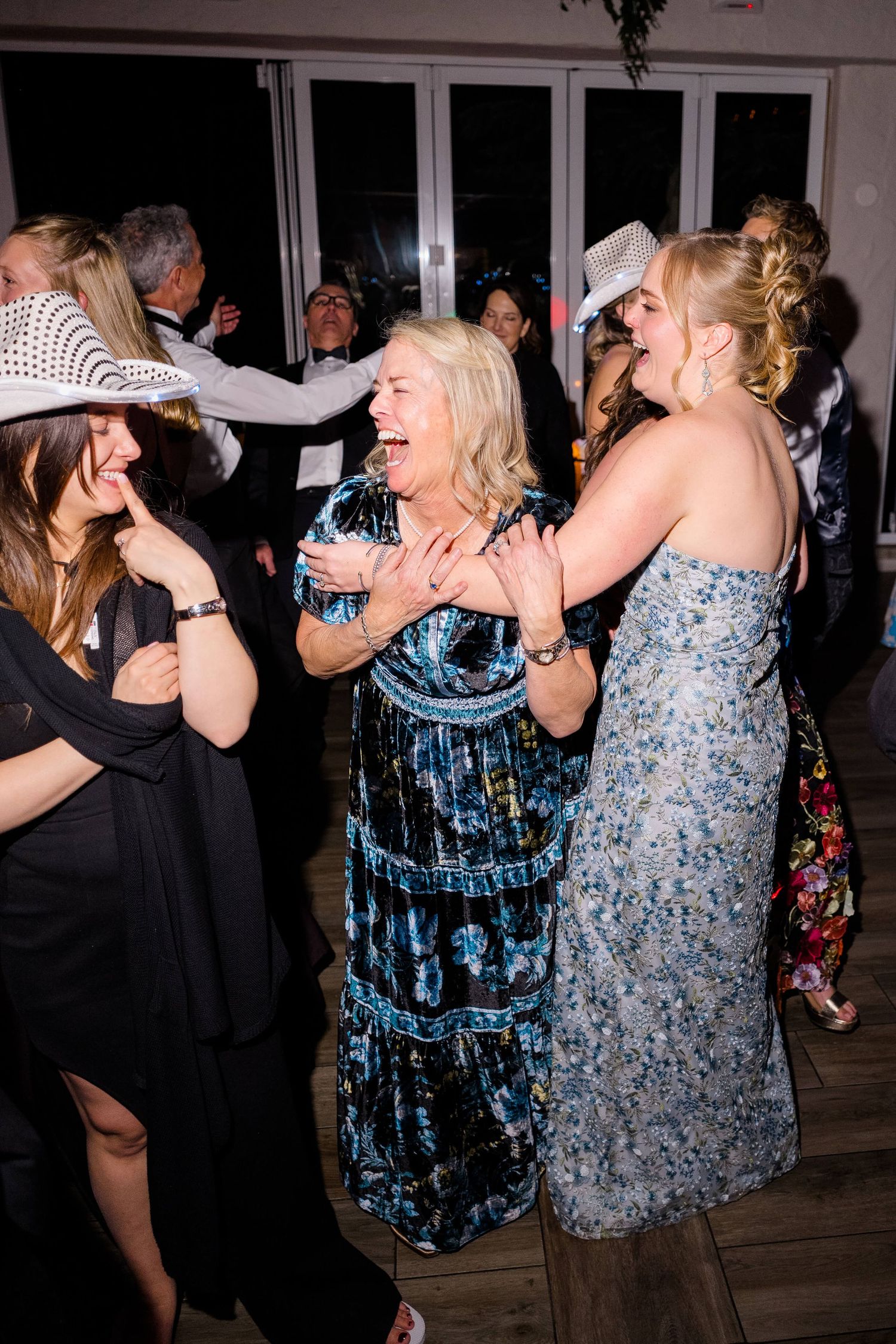 Women laughing and dancing together at a festive party, wearing elegant dresses and white cowboy hats.