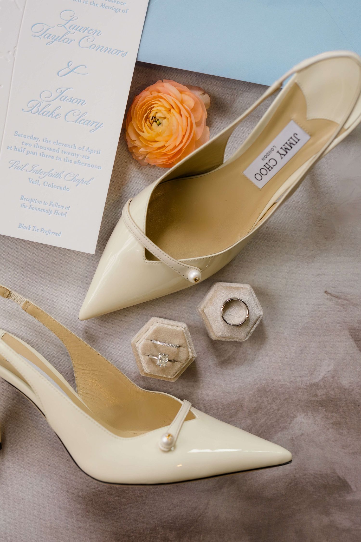 Ivory Jimmy Choo slingback heels with pearl detail beside wedding rings, ranunculus bloom, and blue wedding invitation.