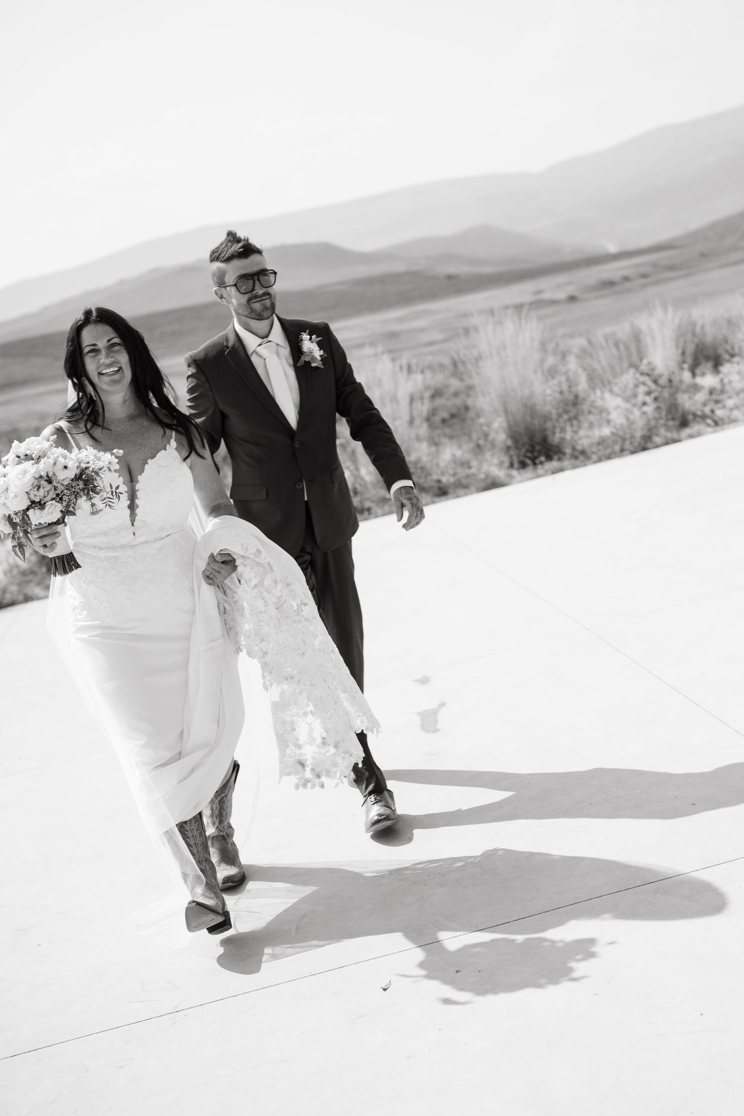 A couple in wedding attire running playfully through snow-covered terrain with mountains in the background.