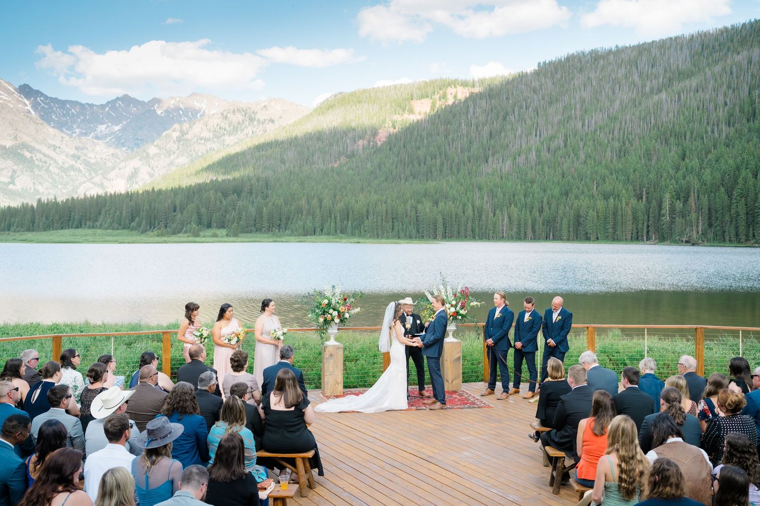 A scenic mountain wedding ceremony with guests gathered on a wooden deck overlooking a serene lake and forest landscape at Piney River Ranch