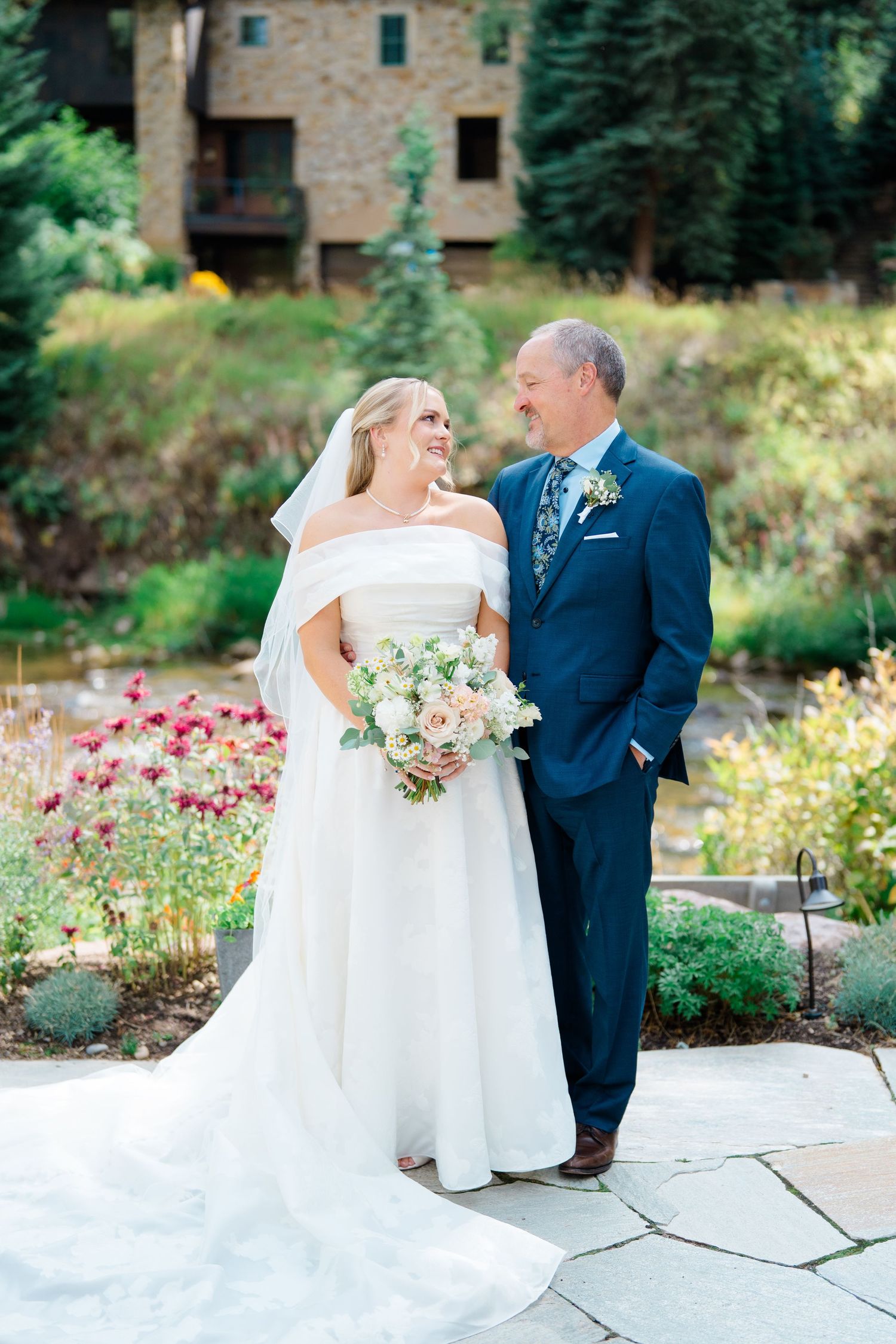 Wedding couple posing in garden with pink flowers and stone patio on sunny afternoon.