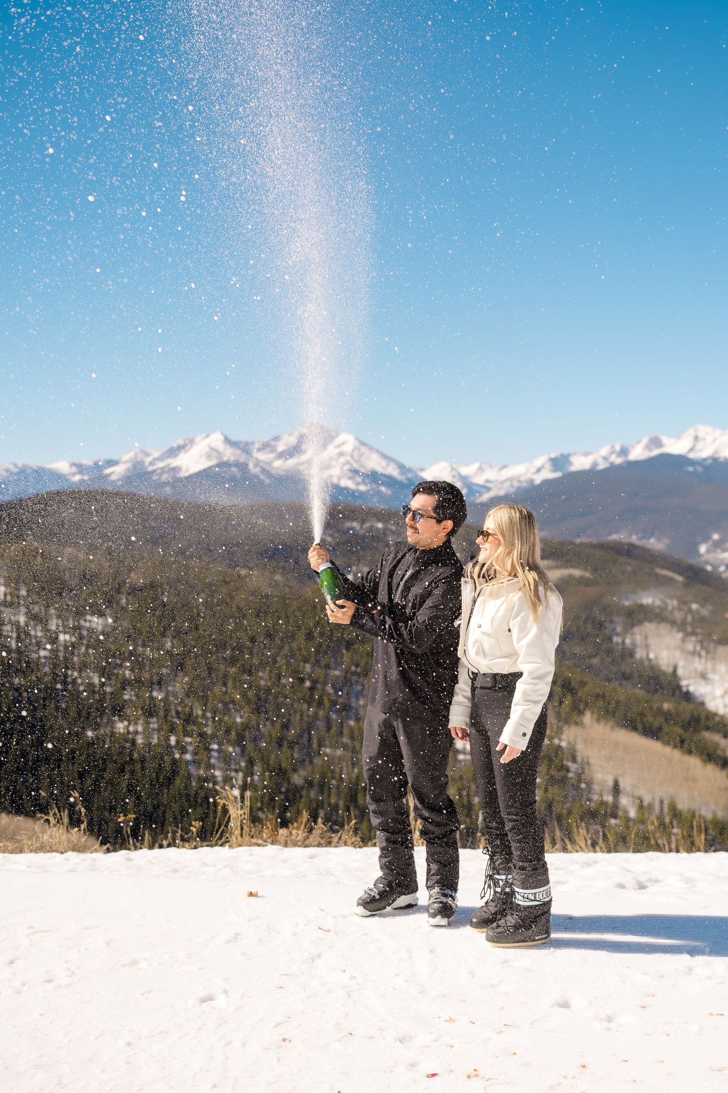 Couple celebrating with champagne on a snowy mountain top with Rocky Mountain peaks in the background.