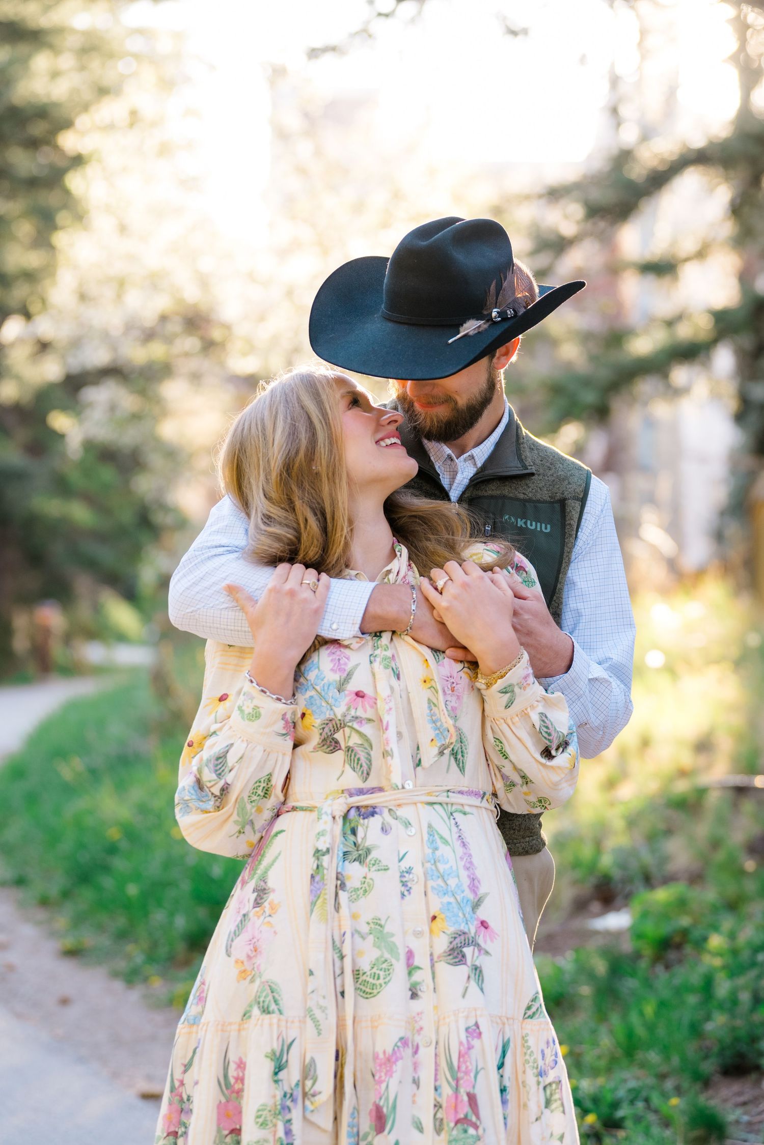 A couple embraces outdoors, woman in floral dress, man in cowboy hat and vest, sharing a romantic moment.