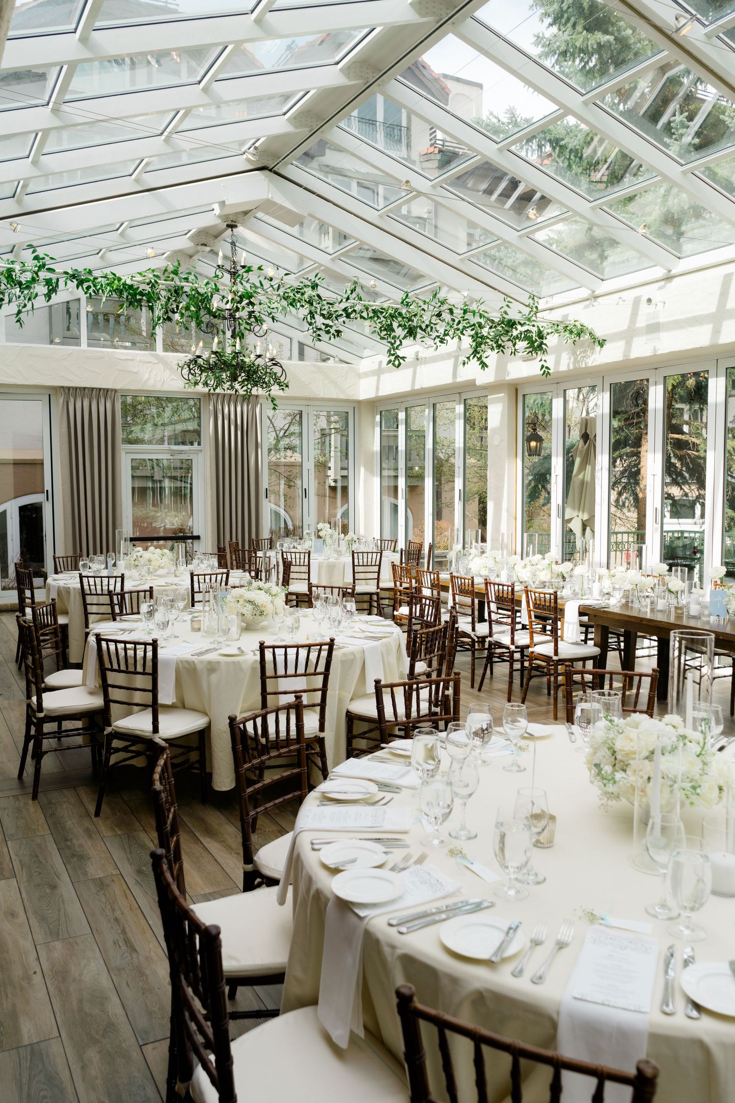 Elegant wedding reception in a glass conservatory with white linens, chiavari chairs, and greenery chandeliers at Vail Sonnenalp Wedding.