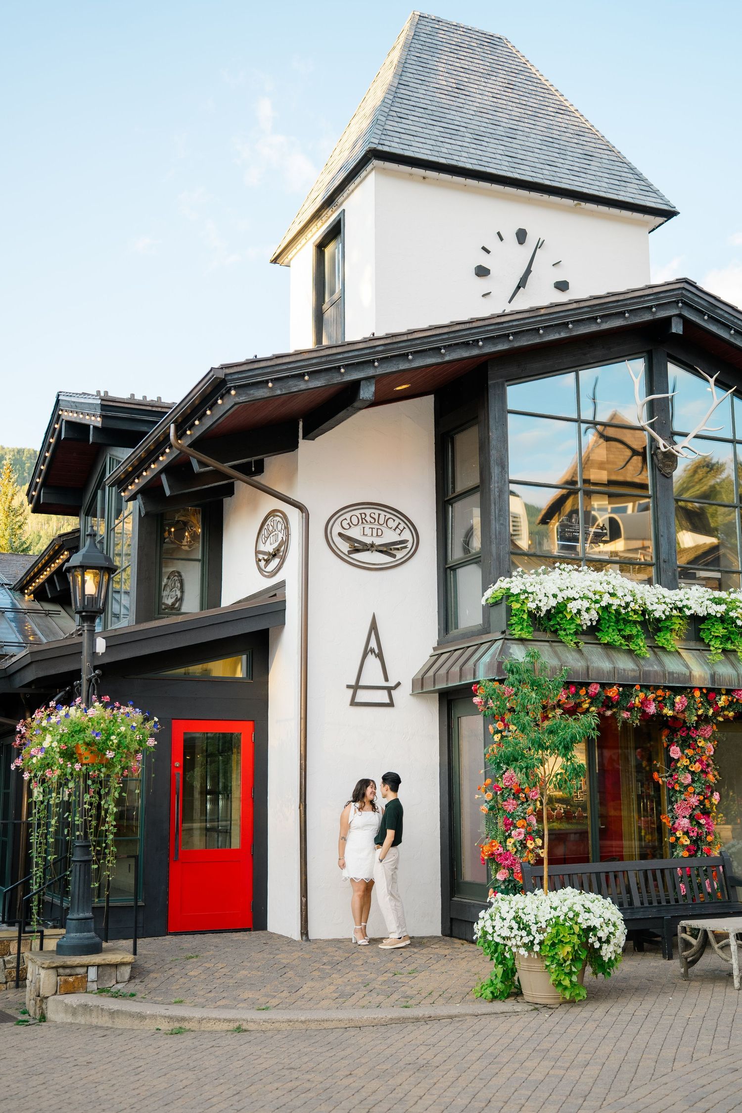 A charming alpine-style building with a clock tower, red door and hanging flower baskets in a mountain village.