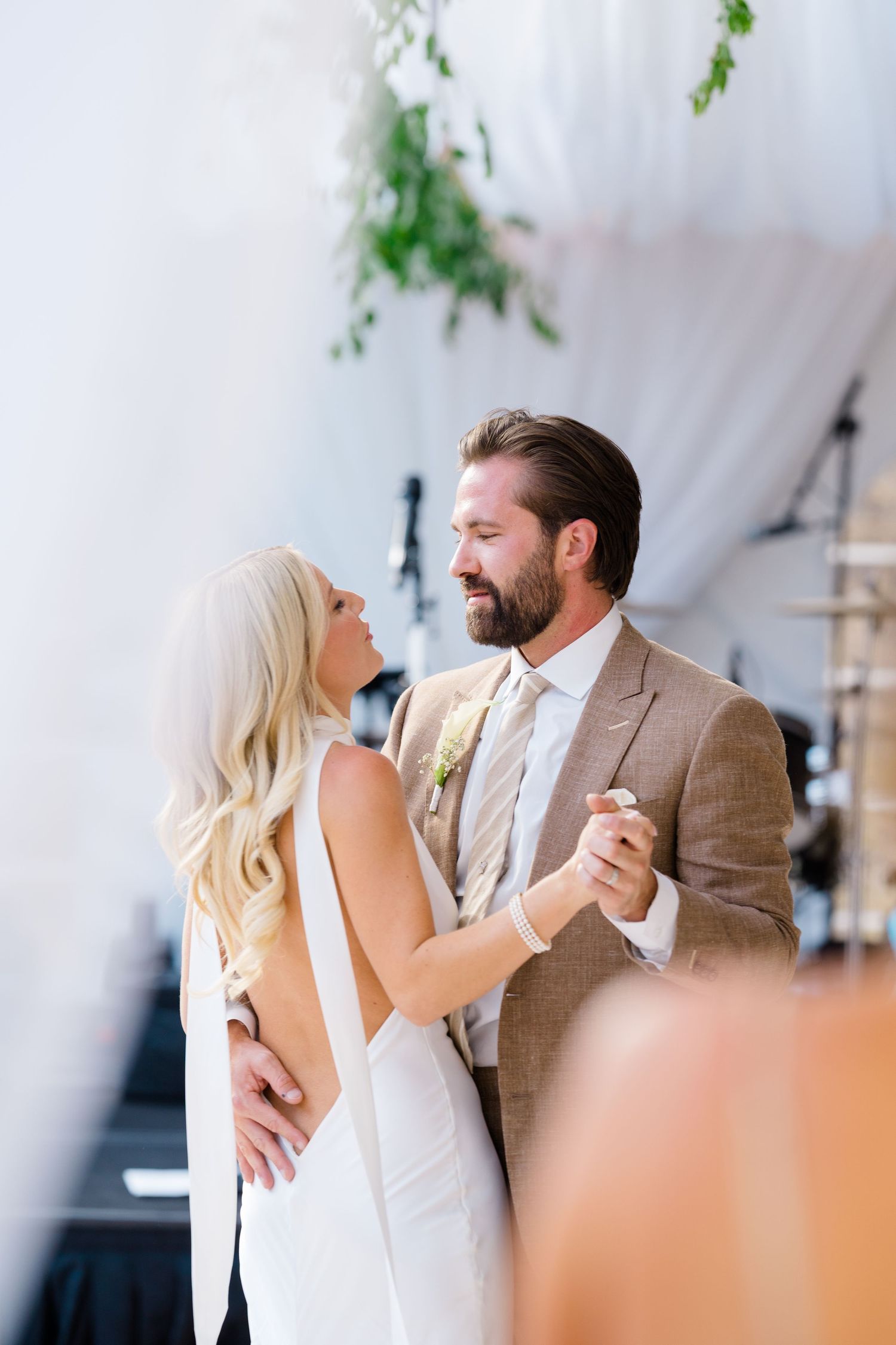 Bride and groom share their first dance, she in white gown, he in tan suit with boutonniere, at elegant wedding reception.