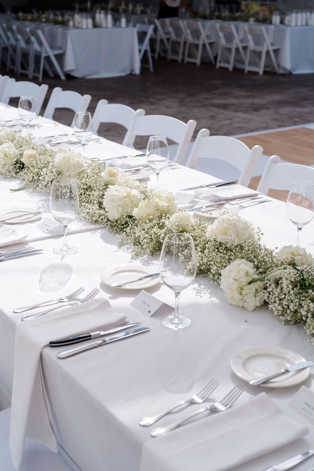 Elegant wedding reception table with white floral runner, baby's breath, wine glasses, and silverware on white linen.