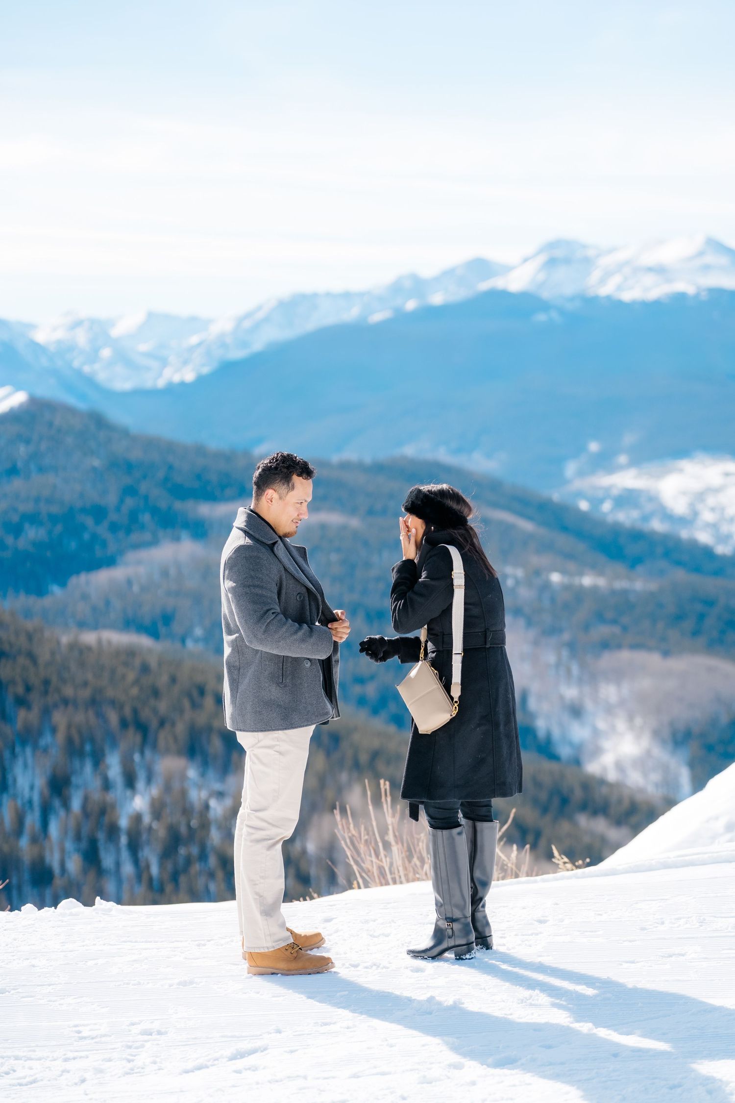 Two people in winter clothing kneel in snow with snowy mountain peaks in background during a proposal moment.