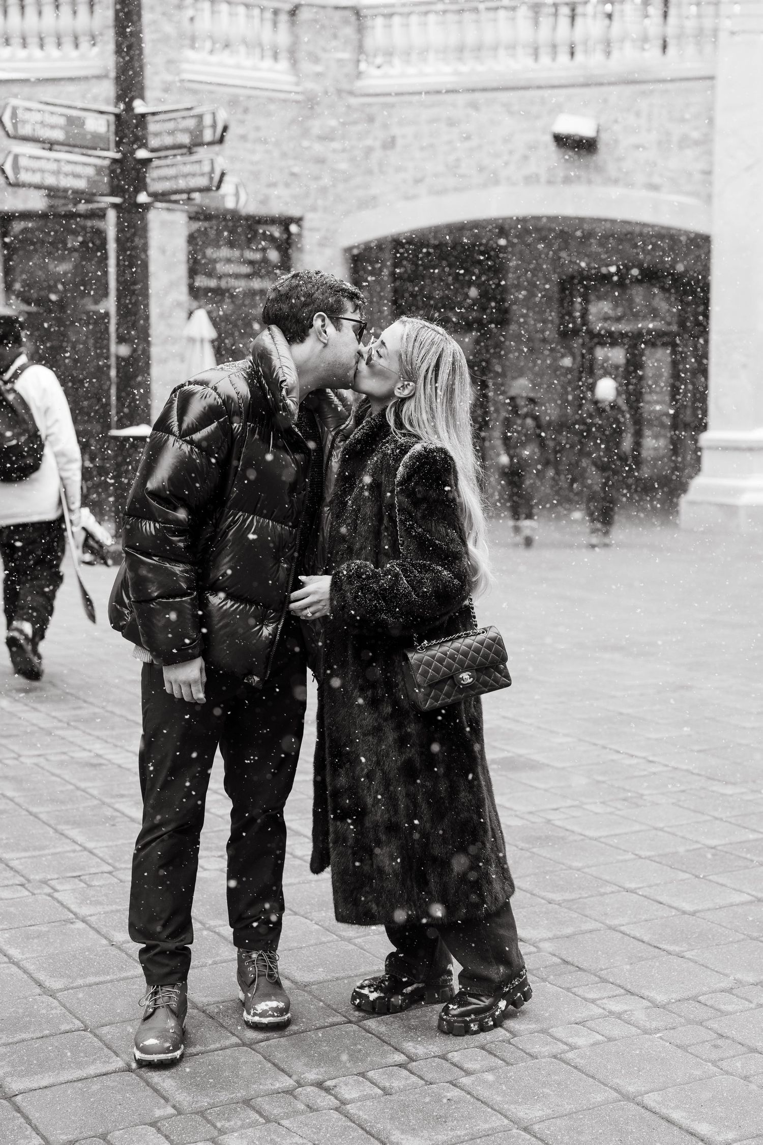 Black and white photo of a couple kissing in the snow on a city street, both wearing winter coats.