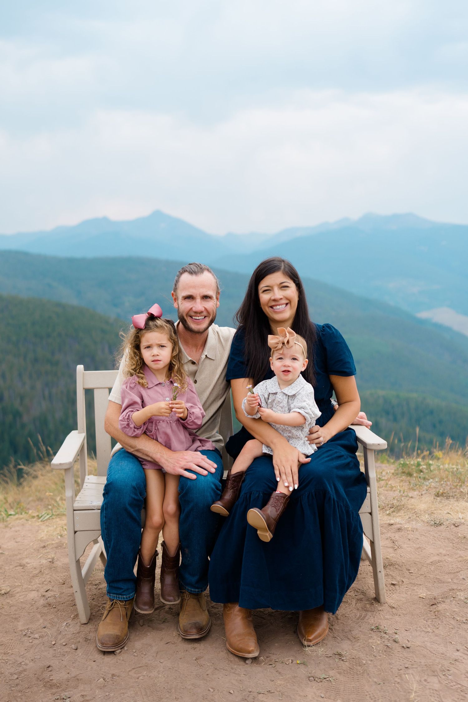 Family sitting together on wooden bench against backdrop of scenic mountain ranges on sunny day during vail mountaintop family photos.