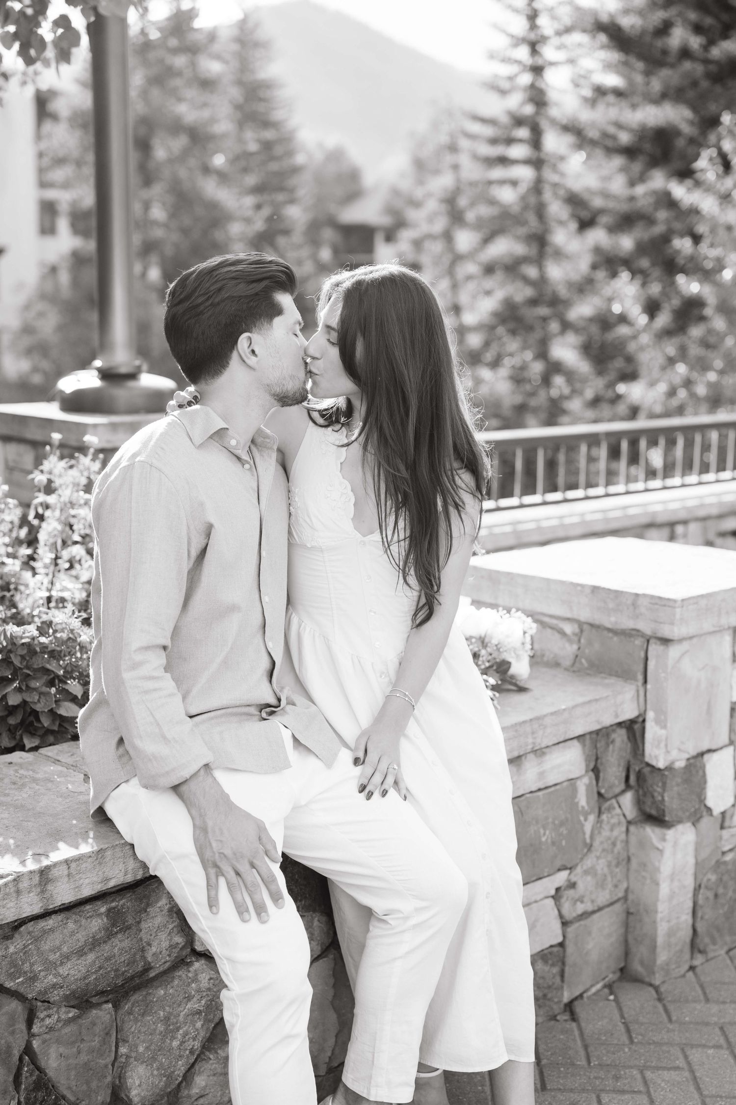 vail village proposal with a romantic couple in white outfits share intimate moments on a stone patio surrounded by evergreen trees.