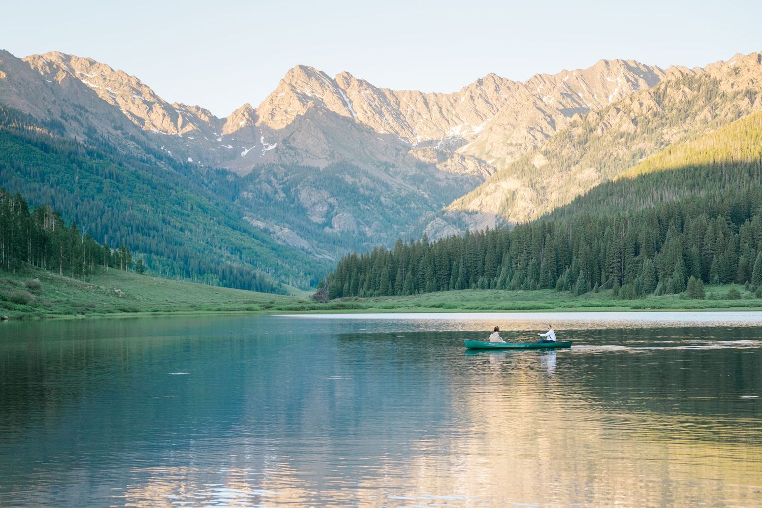 Scenic mountain lake with boat dock surrounded by pine forest and snow-capped peaks reflecting in calm waters at sunset.