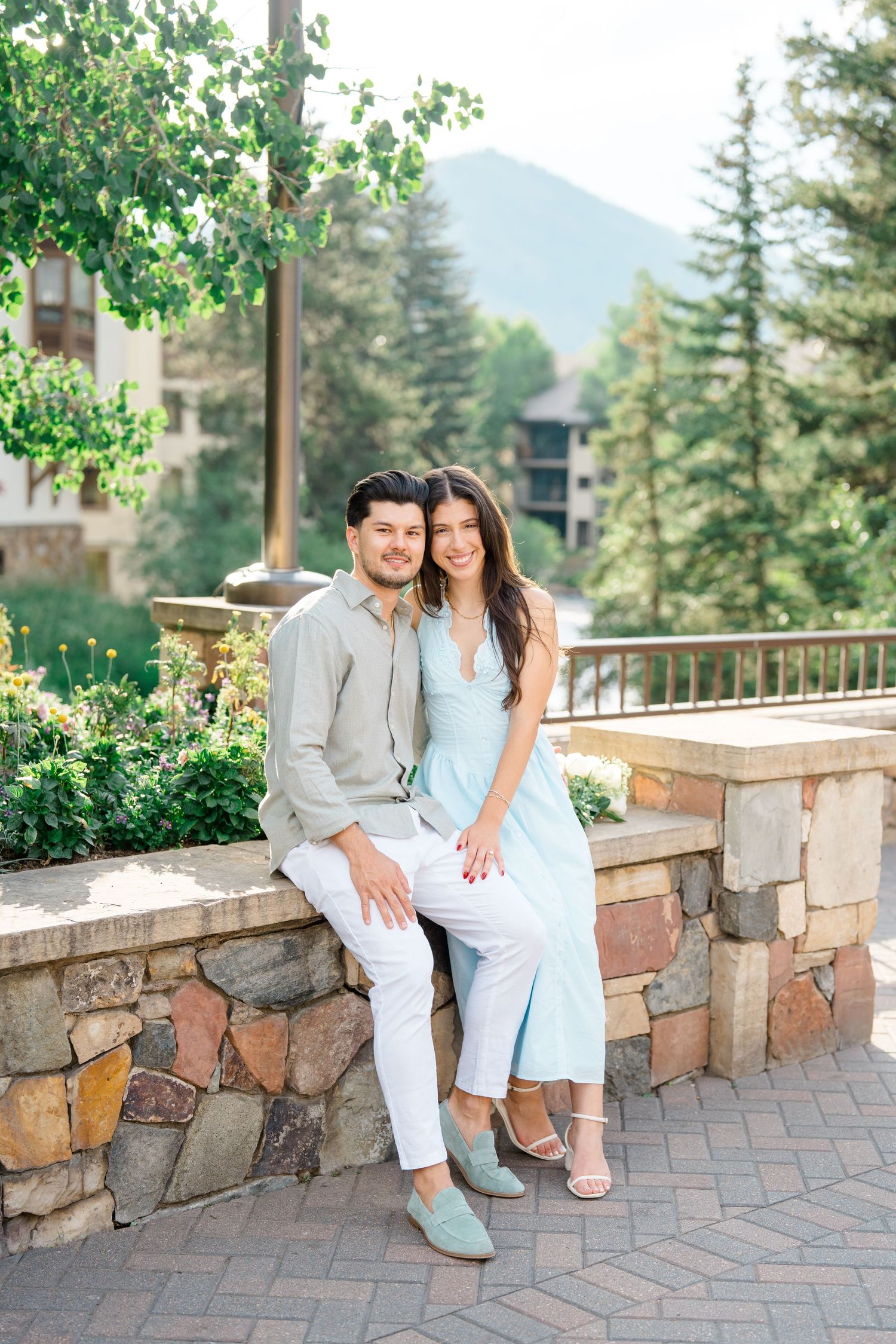 A couple sits together on a stone wall overlooking mountain scenery on a sunny summer day.