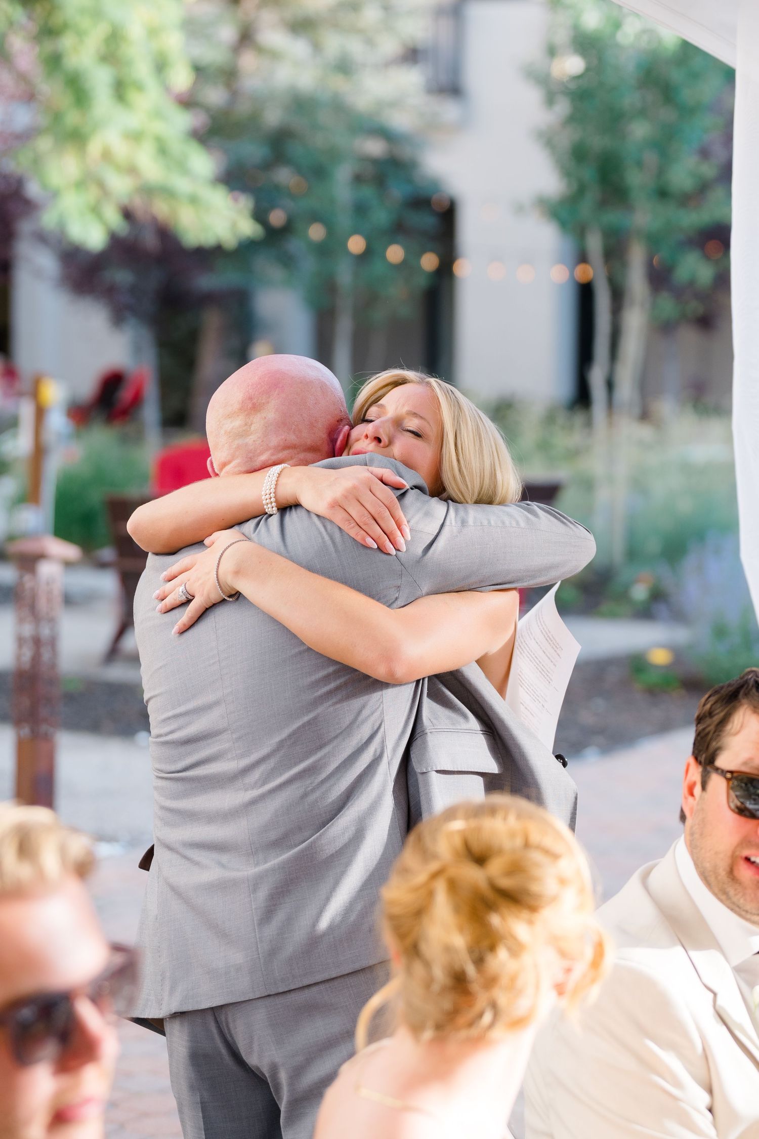 A bride and groom share a warm embrace at an outdoor wedding reception, surrounded by guests and string lights.