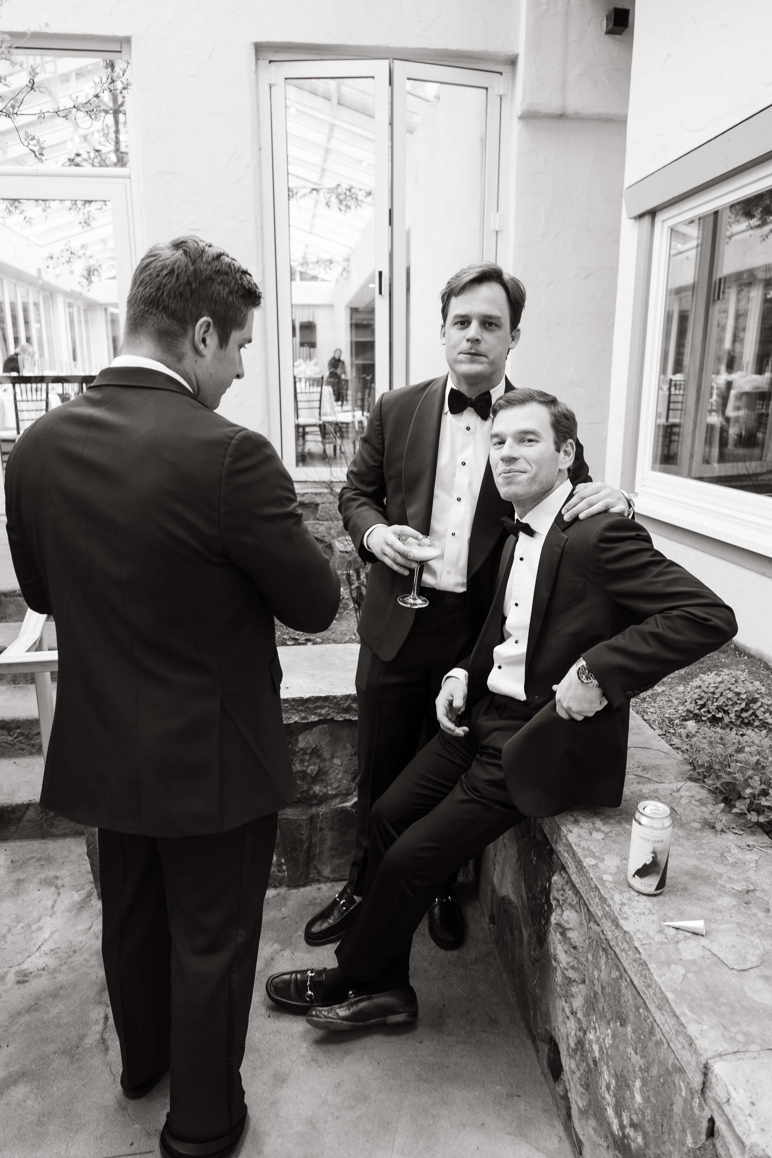 Three men in tuxedos socializing outdoors near a building entrance in a black and white wedding photo.