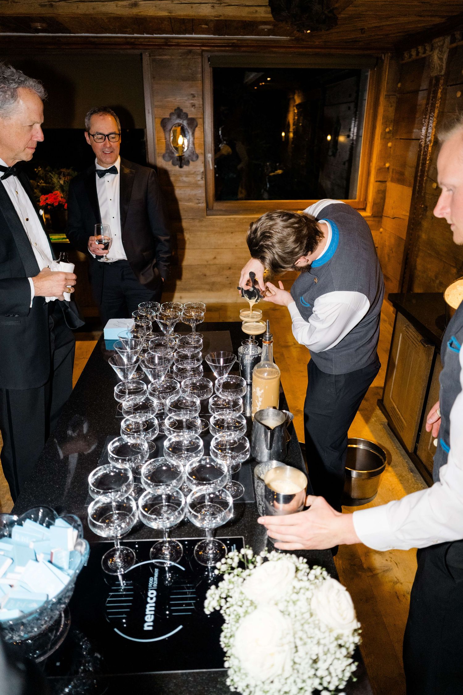 Bartender pouring champagne tower at elegant black-tie event with guests watching in rustic venue.