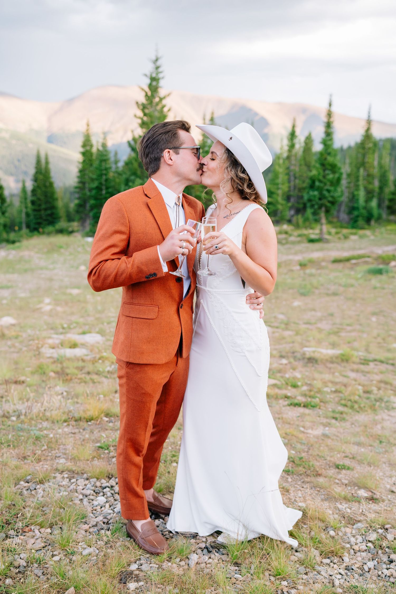 Romantic wedding couple embracing in an orange suit and white dress against a mountain backdrop with evergreen trees at vail wedding.