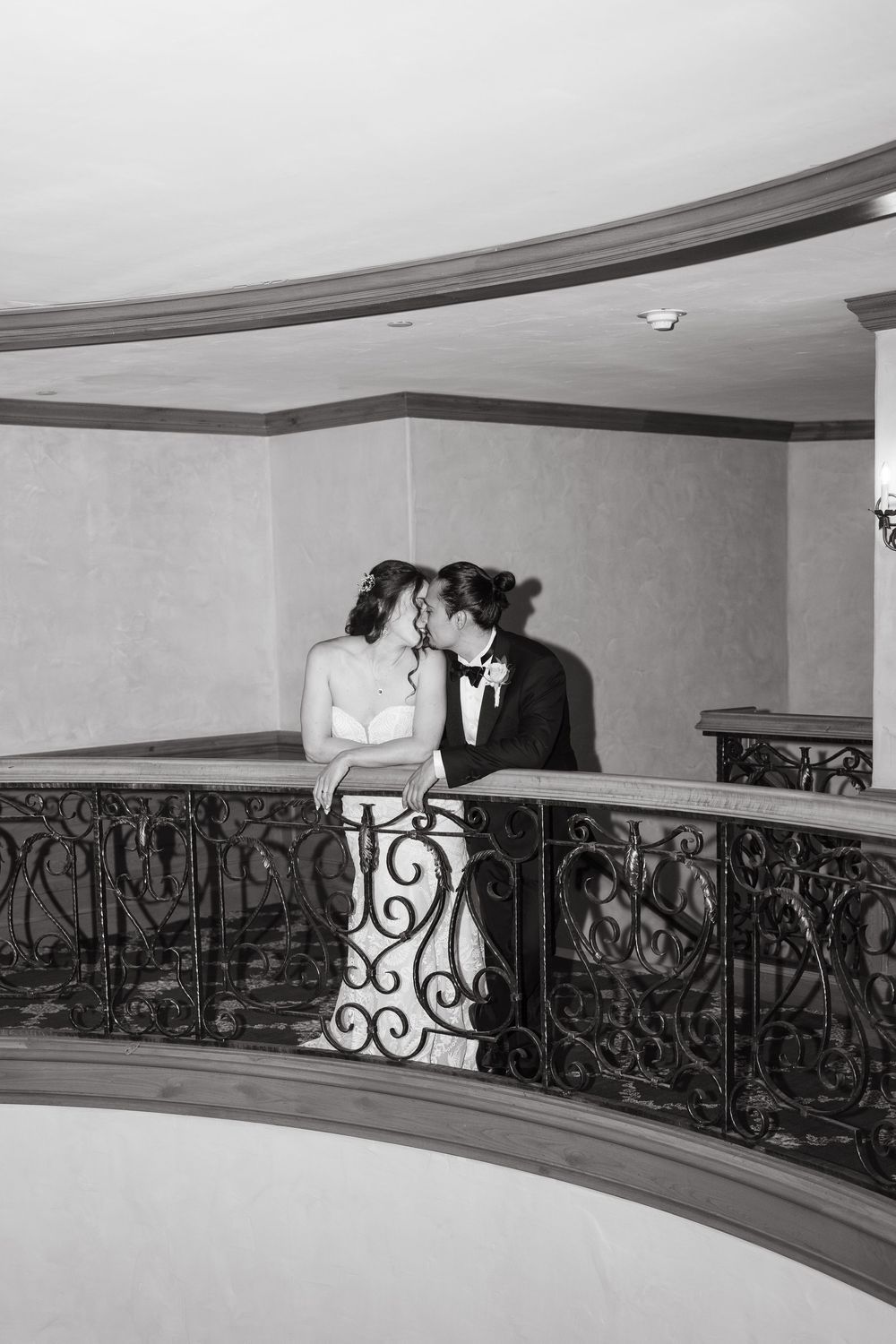 A couple shares a romantic moment along an elegant curved balcony with ornate wrought iron railings in black and white during breck wedding.