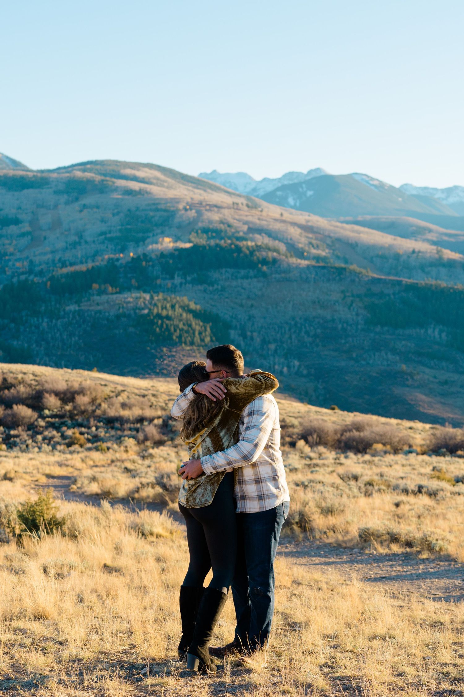 Two people embrace while looking out at mountain vistas and golden grasslands in late afternoon light.