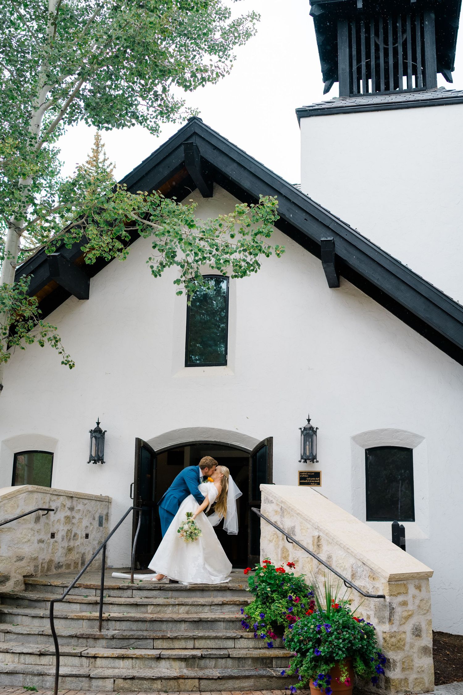 Wedding couple kisses on church steps decorated with floral arrangements and lanterns.