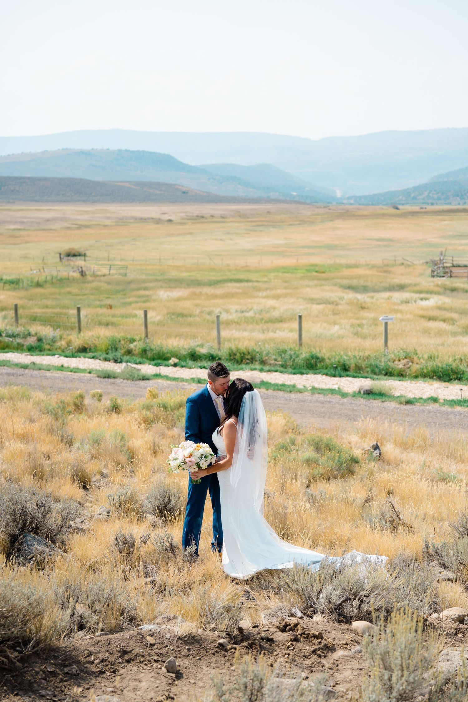 A couple shares an intimate moment in a rustic field with mountains and golden grass in the background.