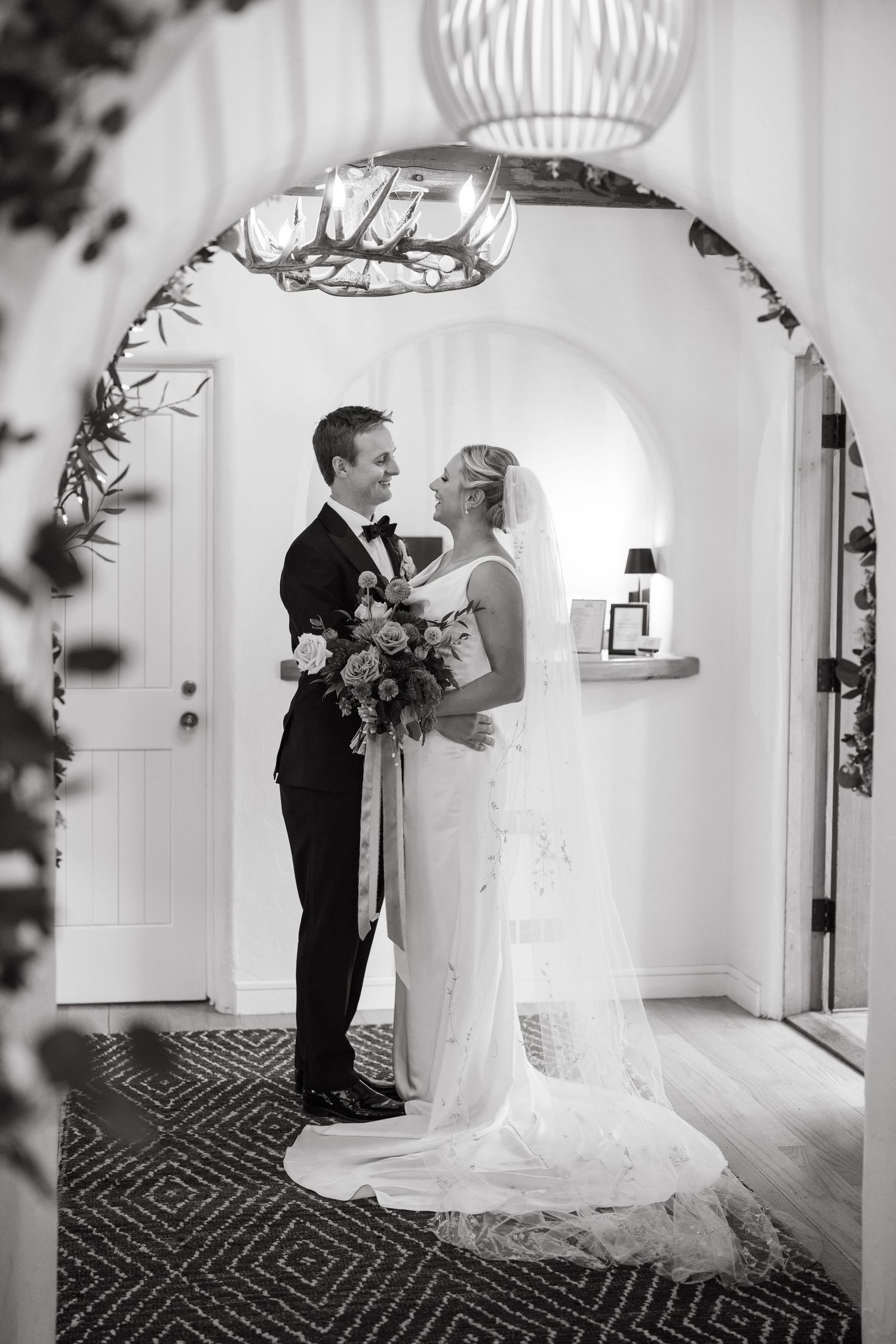 A romantic black and white wedding photo of a couple standing in an elegant arched doorway with decorative lighting.
