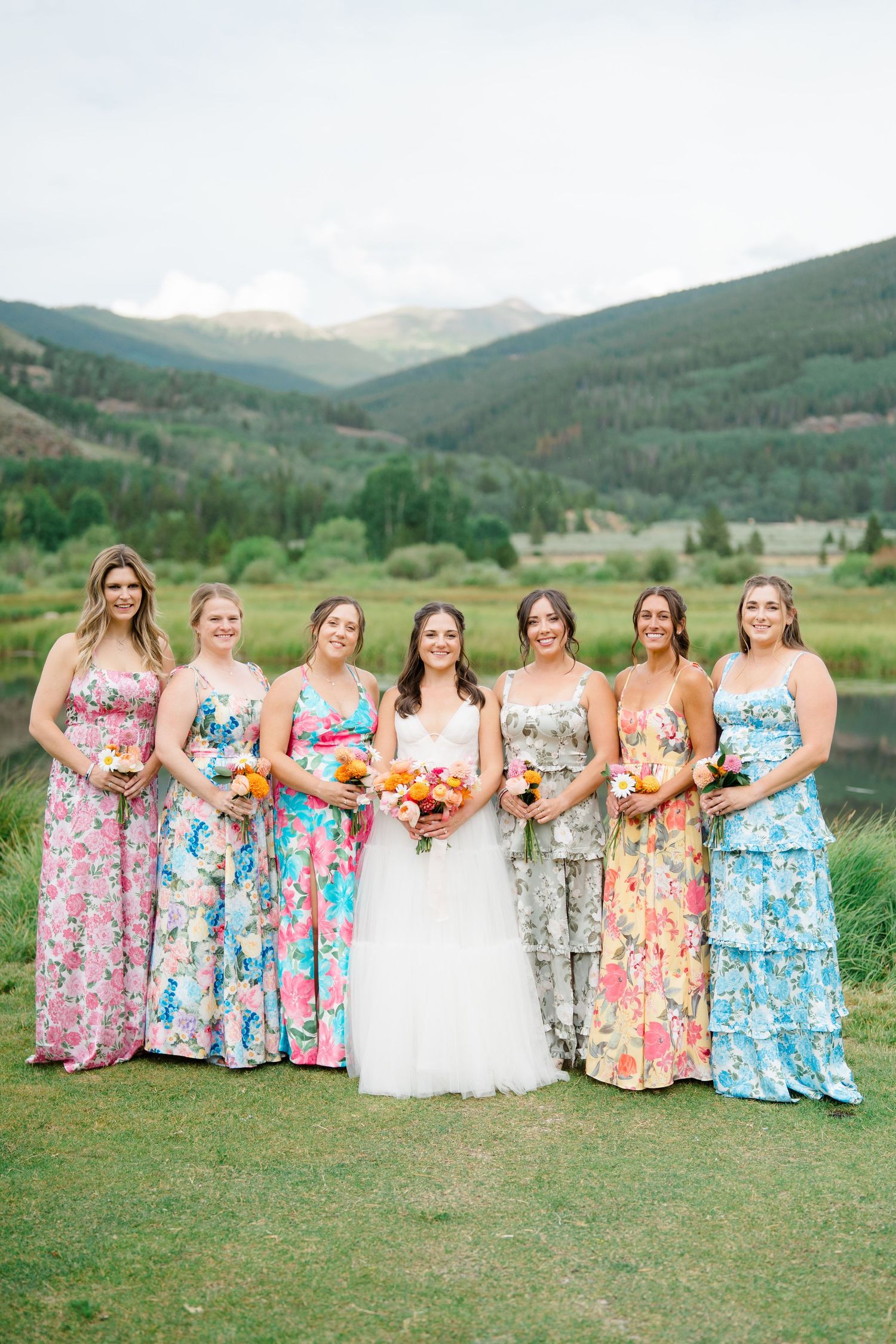 Wedding party in colorful floral dresses posing in a mountain meadow setting.