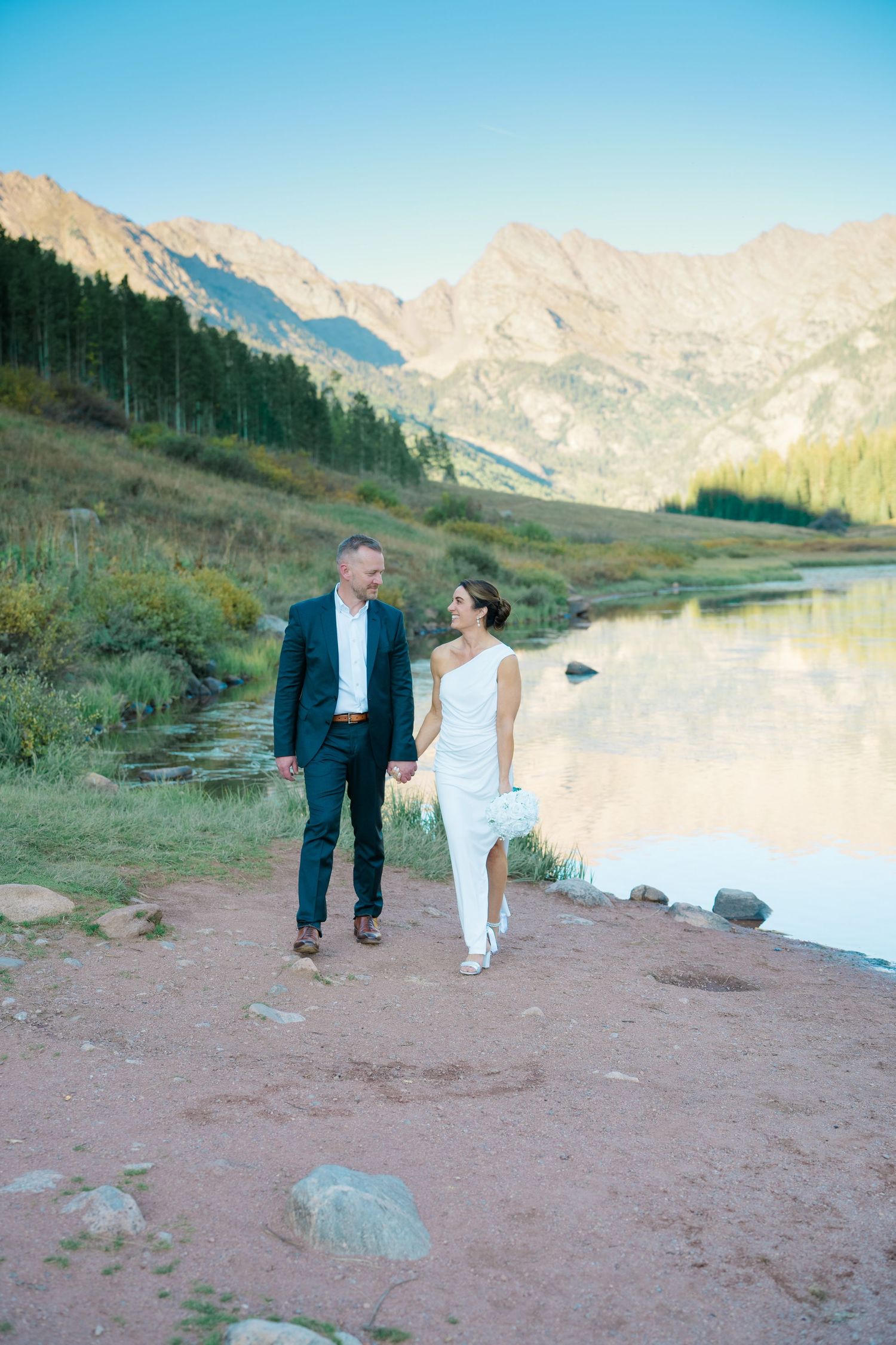 Couple walking together along a mountain lake shoreline at sunset with rocky peaks in the background during piney river ranch elopement.