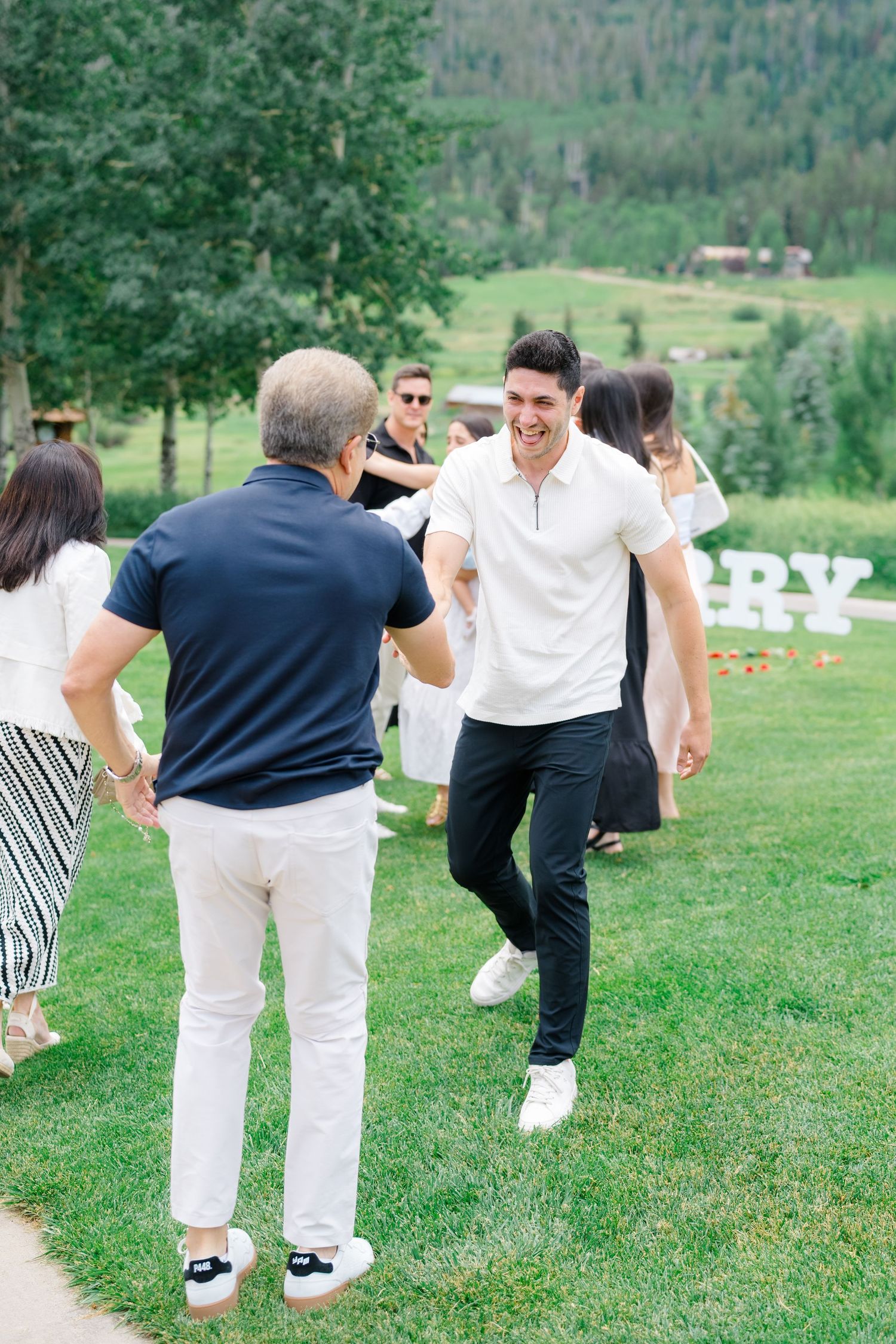 People in white and navy outfits playing games on a grassy hillside with mountain views.