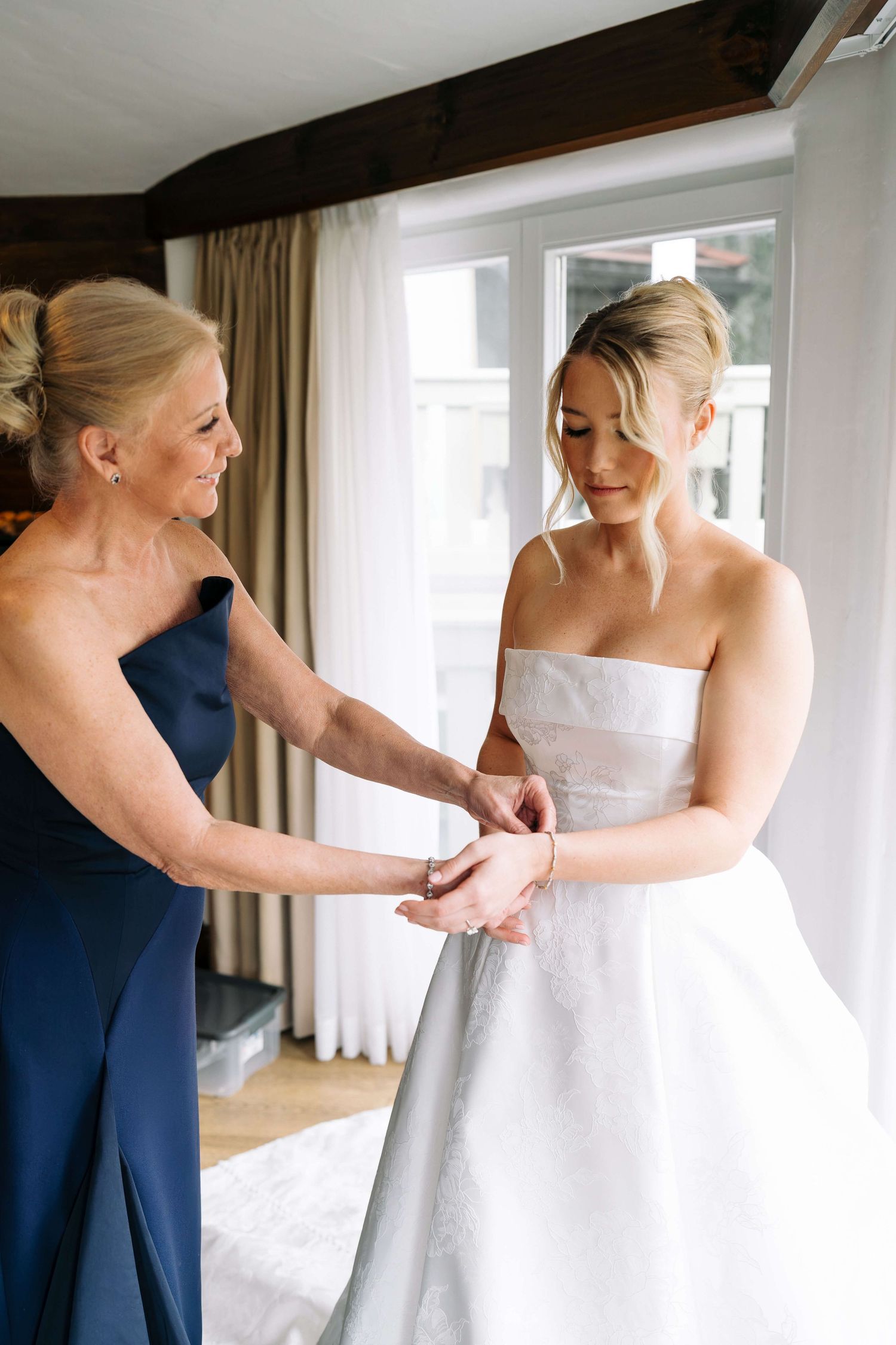 Mother in navy blue dress helping bride in strapless white wedding gown with final preparations near a bright window.